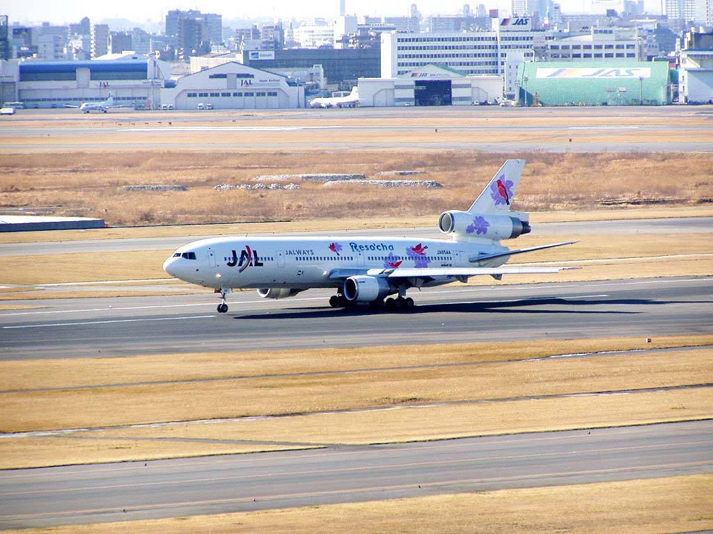 日本 桌布 壁紙 — 一架白色JAL Reso'cha塗裝的波音767在陽光明媚的機場跑道上滑行，背景有機庫和城市建築。
