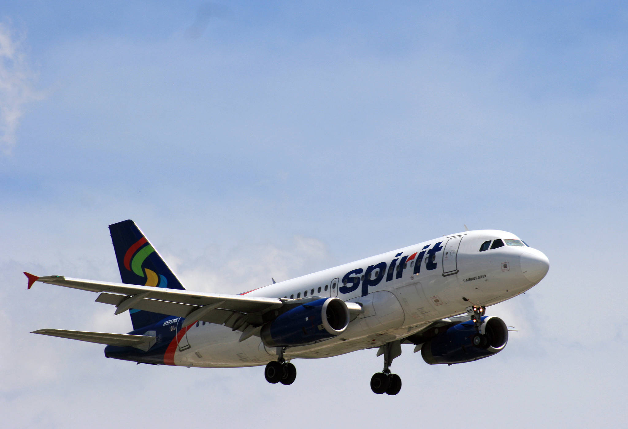 Blue Sky 桌布 壁紙 — 一架白色Spirit Airlines空中巴士A320，配備藍色引擎和色彩繽紛的尾翼，正對著淡藍色天空和零星雲朵下降。