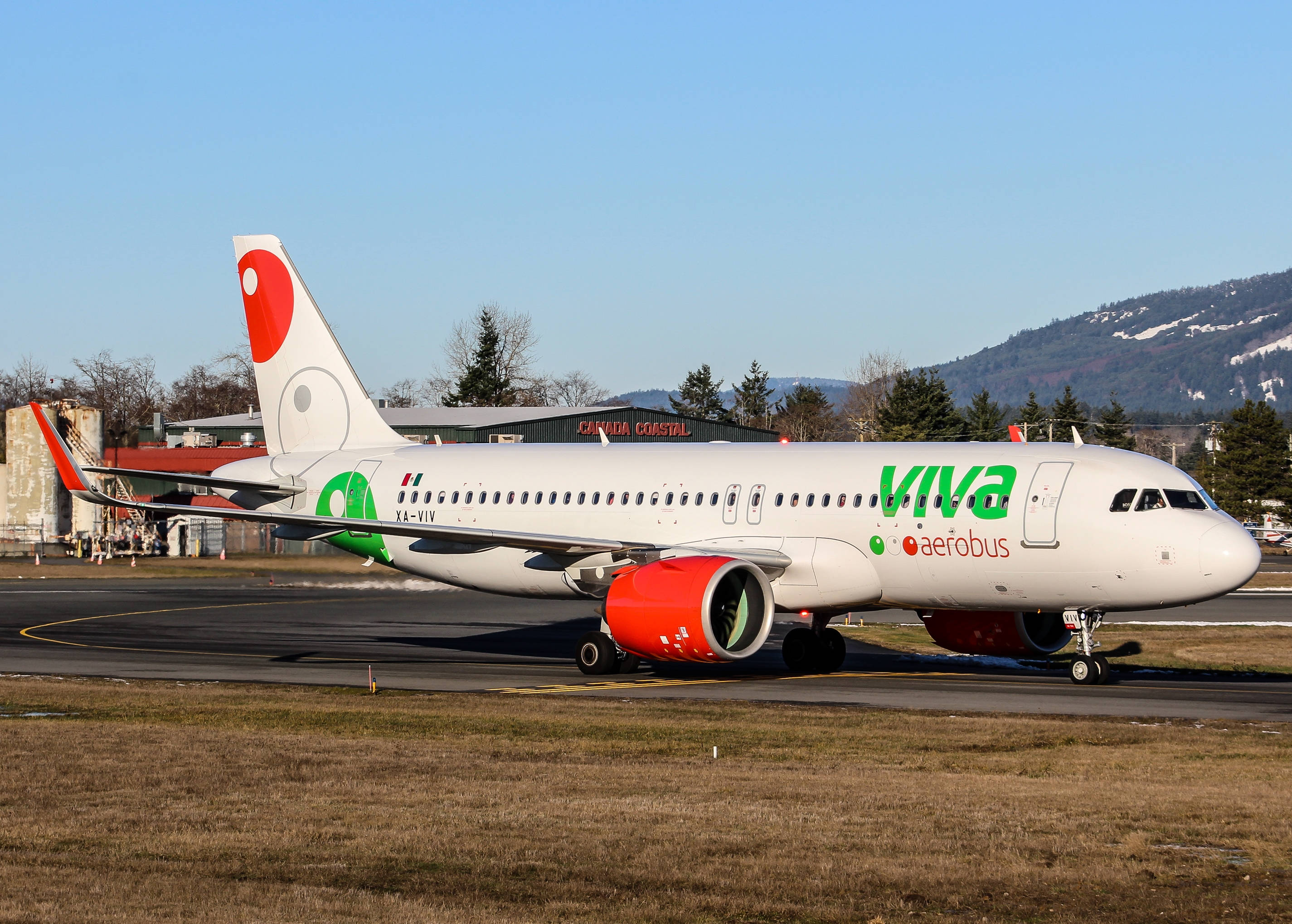 飛機 桌布 壁紙 — 一架白色Viva Aerobus飛機配備紅色引擎，在晴朗藍天和遠處積雪覆蓋的山脈背景下在跑道上滑行。