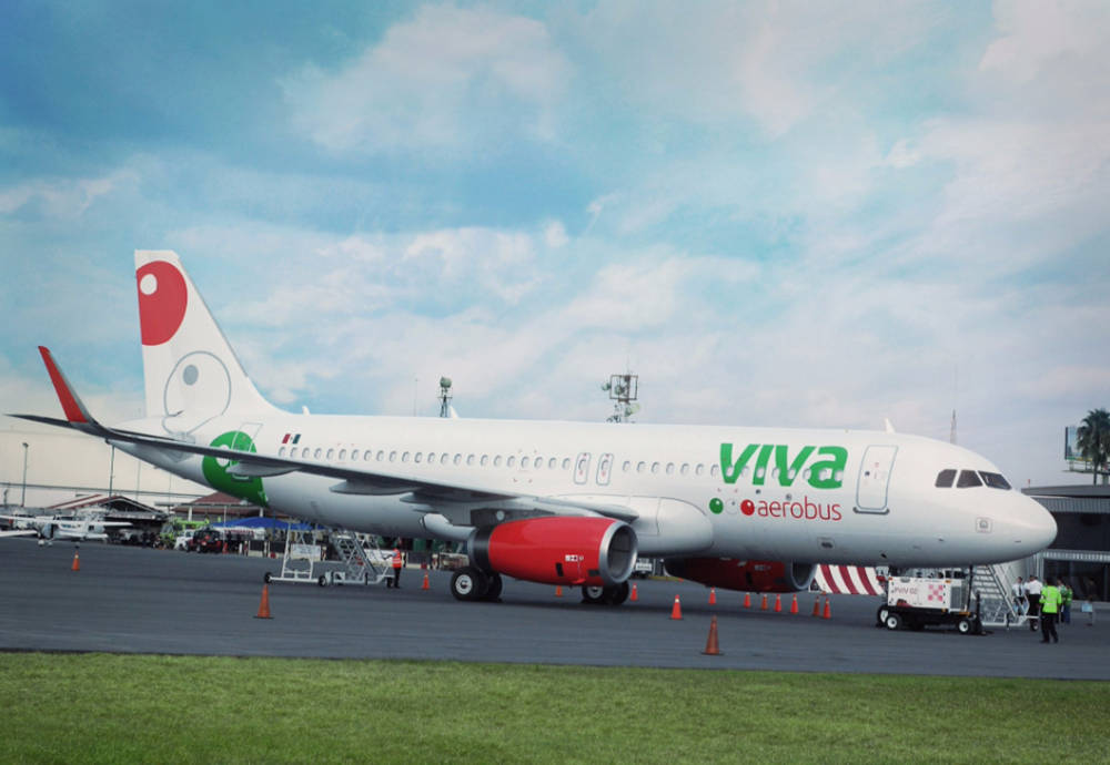 Grass 桌布 壁紙 — 一架白色Viva Aerobus飛機，配備紅色引擎和綠色品牌標識，停在部分多雲天空下的機場停機坪上。