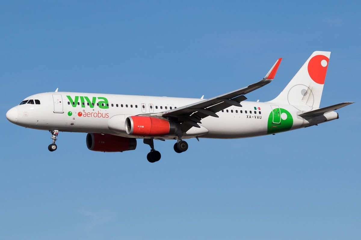 飛機 桌布 壁紙 — 一架白色Viva Aerobus飛機，紅色引擎和綠色品牌標誌，在晴朗的藍天下放下起落架飛行。
