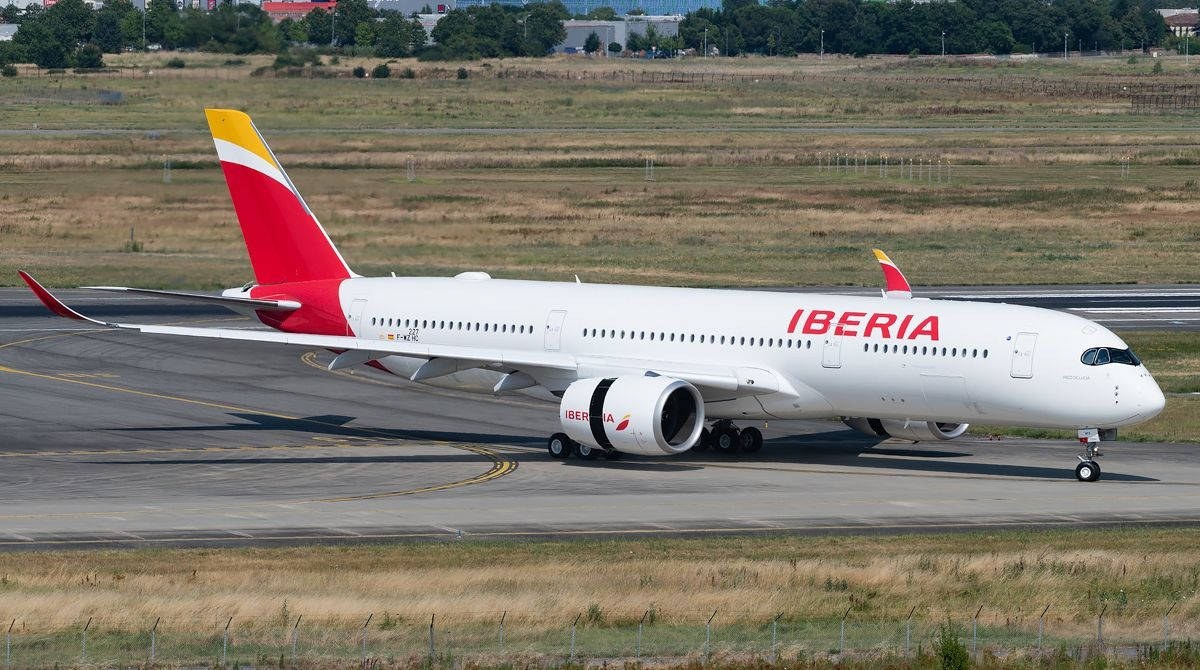 飛機 桌布 壁紙 — 一架白色伊比利亞航空空中巴士A350，尾翼紅黃相間，在明亮的日光下滑行於被乾草和遠方樹木環繞的跑道上。
