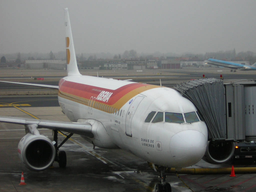 飛機 桌布 壁紙 — 一架白色伊比利亞航空飛機帶有紅色和金色條紋，停泊在濕滑停機坪的登機閘口，天空多雲。
