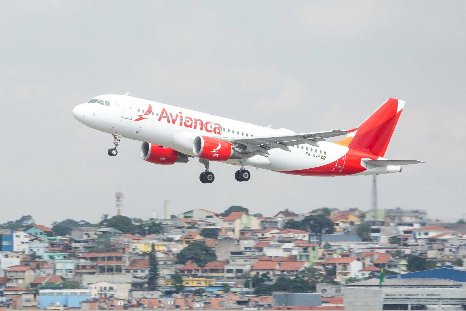 Plane 桌布 壁紙 — 一架白色和紅色塗裝的 Avianca 飛機在多雲的天空下從密集的城市社區上方起飛。