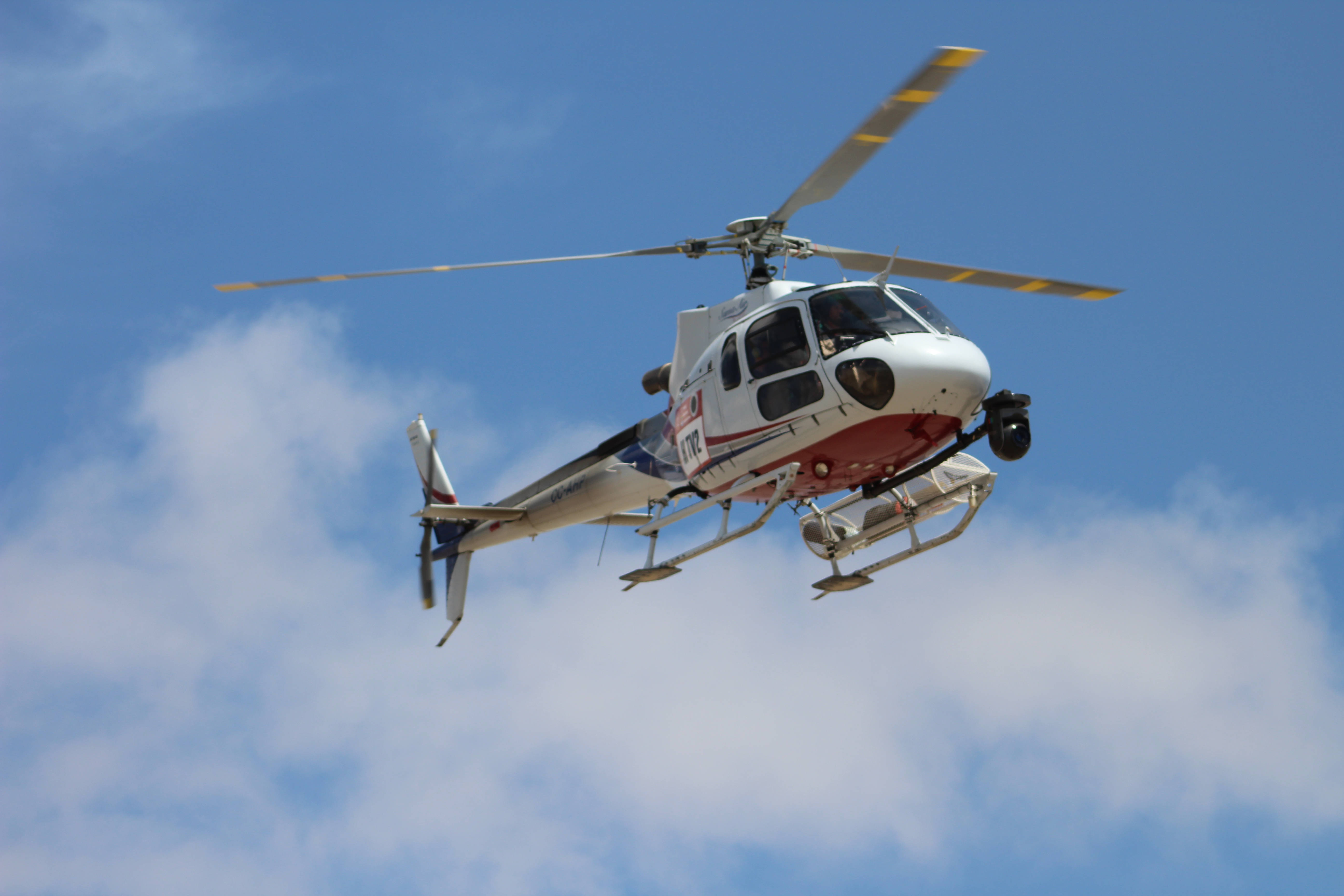 Helicopter 桌布 壁紙 — 一架白色和紅色的直升機，帶有黃色尖端葉片，在明亮的藍天中飛行，天空中有散落的白色雲朵。