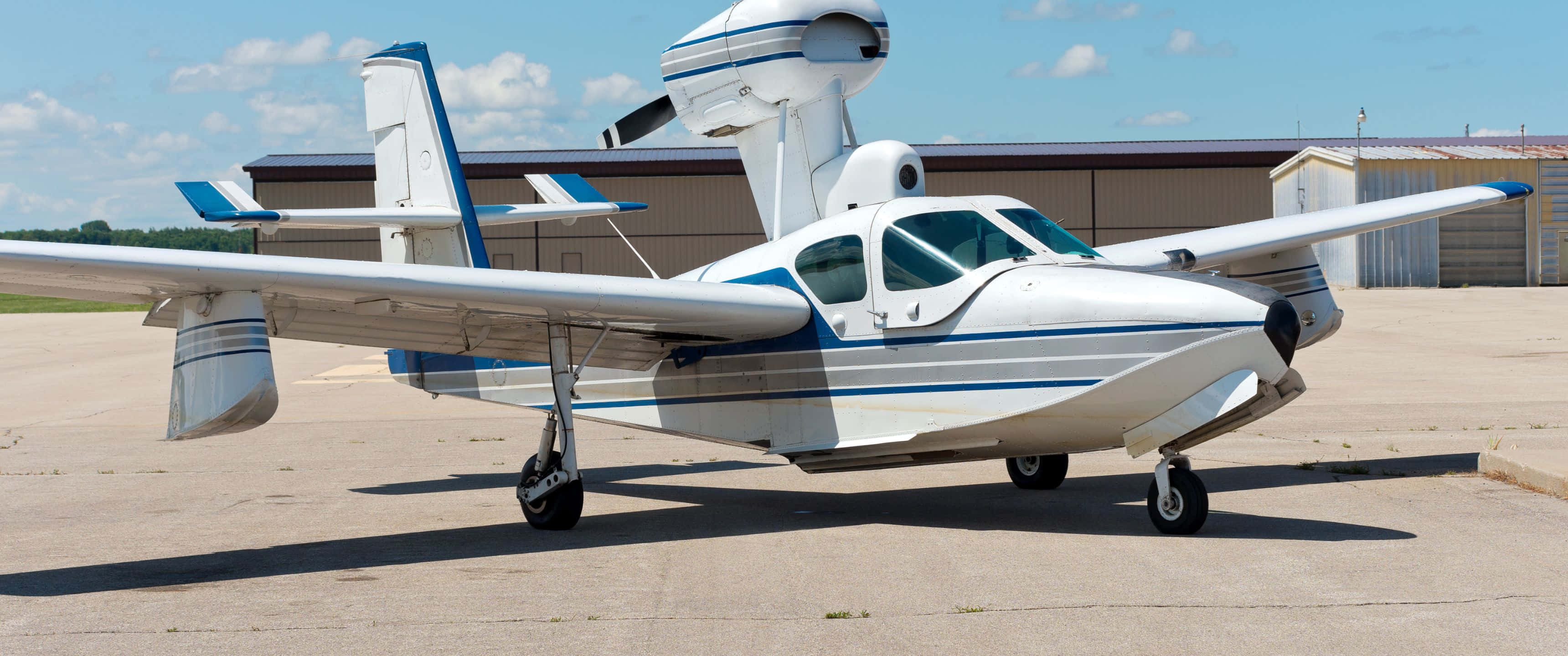 3440x1440p Small Planes 桌布 壁紙 — 一架白色和藍色、帶浮筒的水上飛機停在停機坪上，上方是晴朗的藍天，點綴著幾朵白雲。