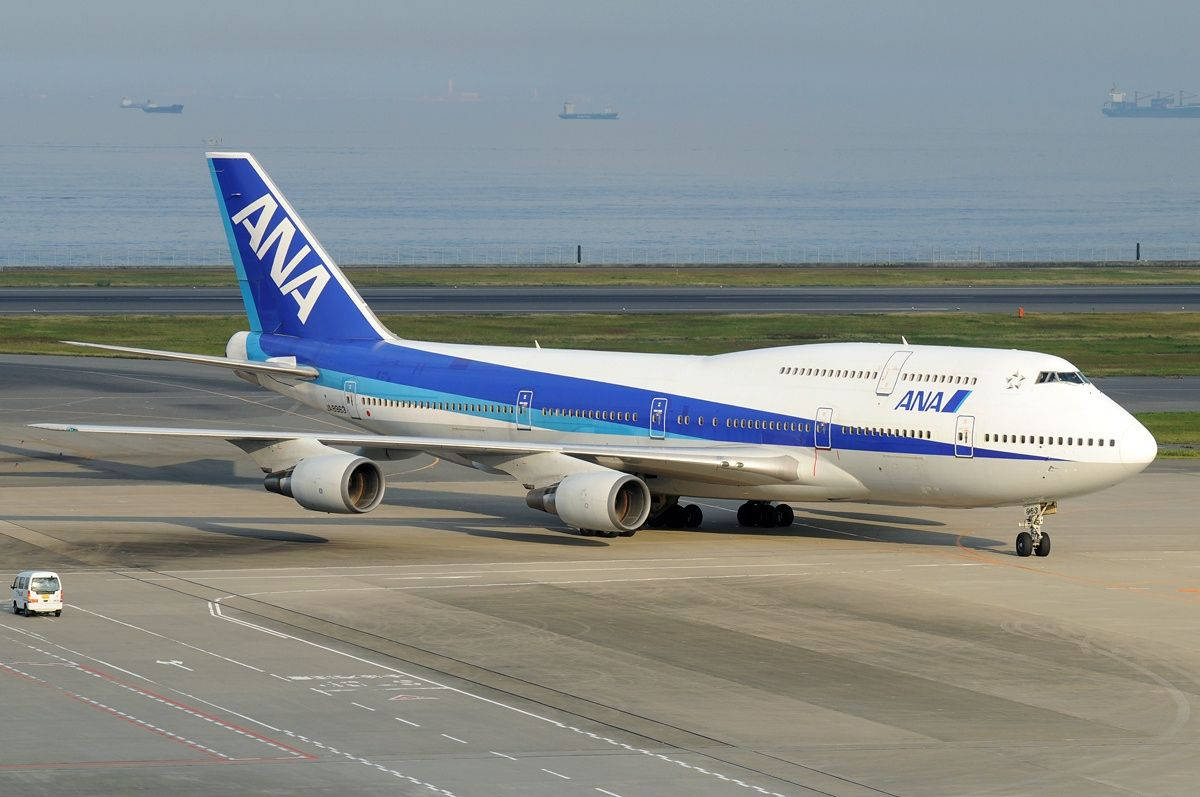 Airport 桌布 壁紙 — 一架白色和藍色的ANA波音747在跑道上滑行，背景是朦朧的海洋和遠處的船隻。
