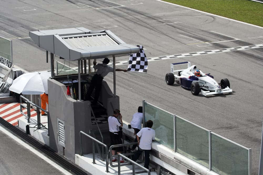 Chequered Flag 桌布 壁紙 — 一架白色和藍色的一級方程式賽車在陽光明媚的賽道上越過終點線，同時一名賽道工作人員揮舞著方格旗。