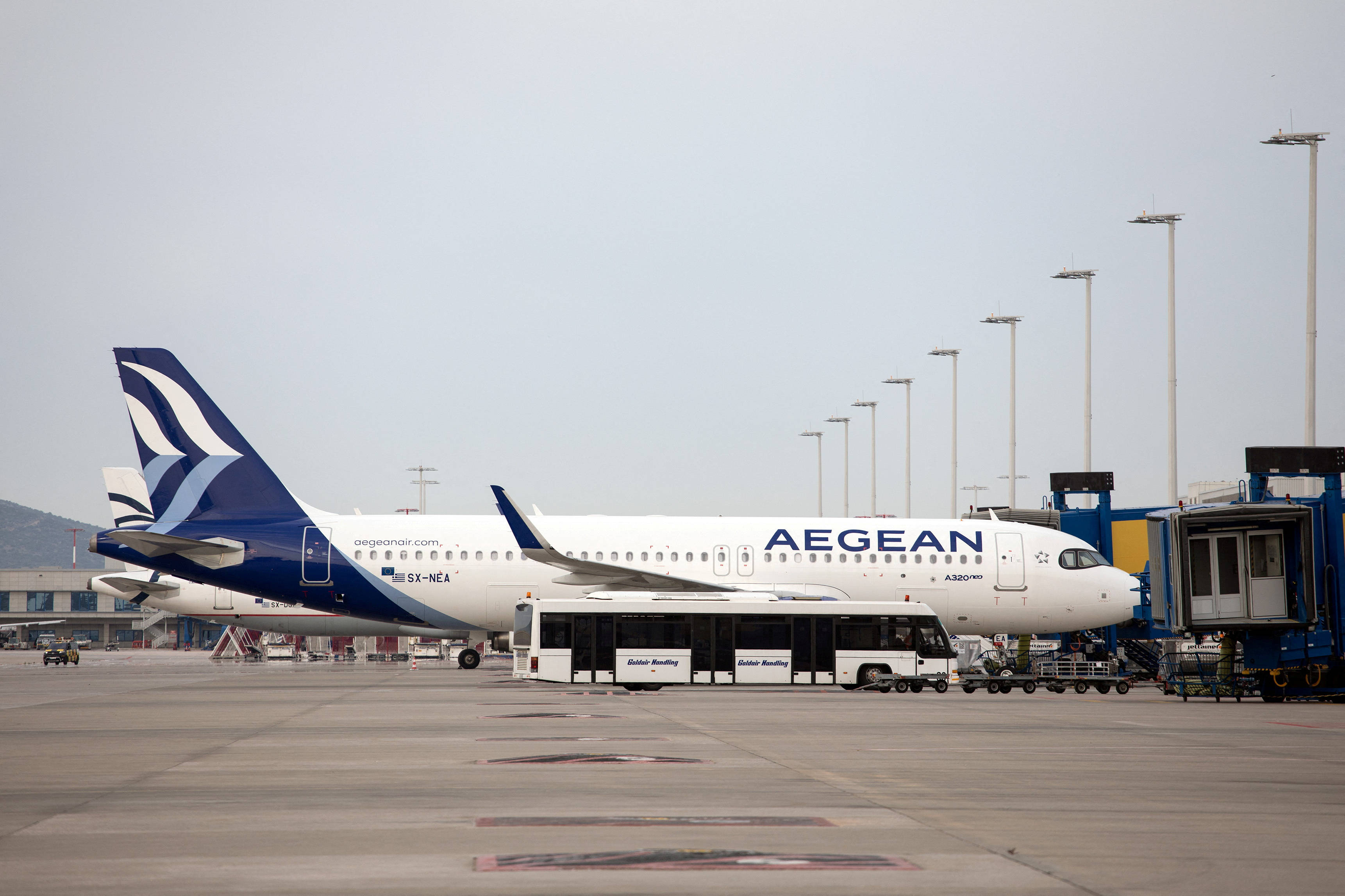 Airport 桌布 壁紙 — 一架白色和藍色的愛琴海航空空中巴士A320停泊在機場閘口，附近有一輛接駁巴士，天空多雲。