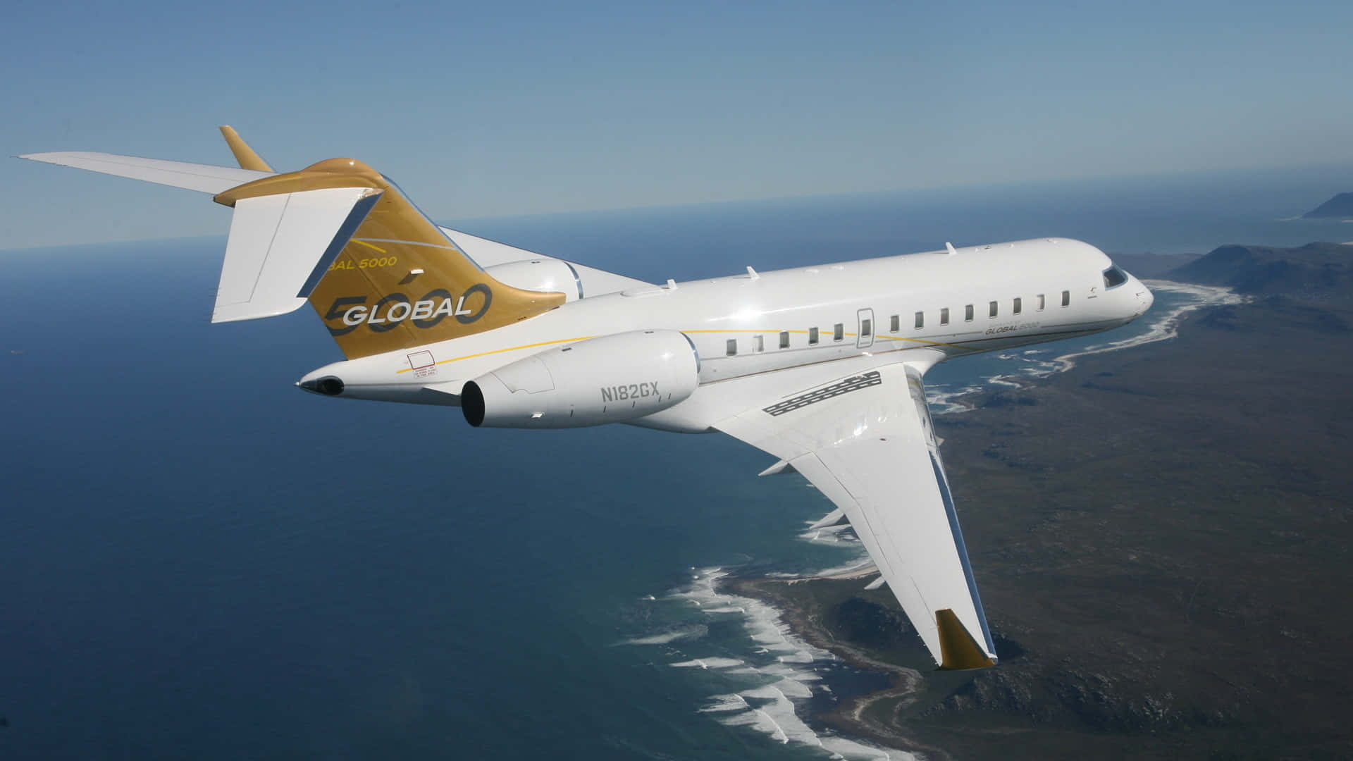 飛機 桌布 壁紙 — 一架白色和金色的Global 6000私人飛機翱翔在深藍色海洋和崎嶇地形的海岸景觀之上。