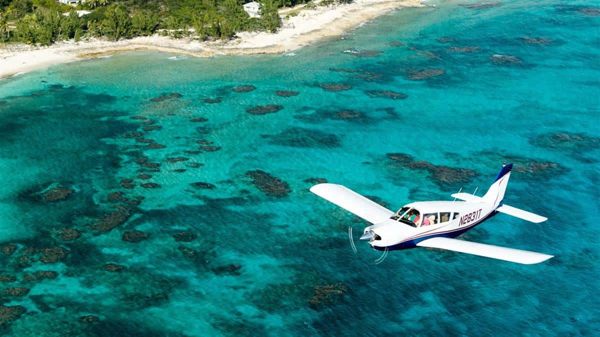 Bahamas Island 桌布 壁紙 — 一架白色單引擎飛機低飛過充滿活力的碧綠海水，露出珊瑚礁和綠樹成蔭的沙灘。