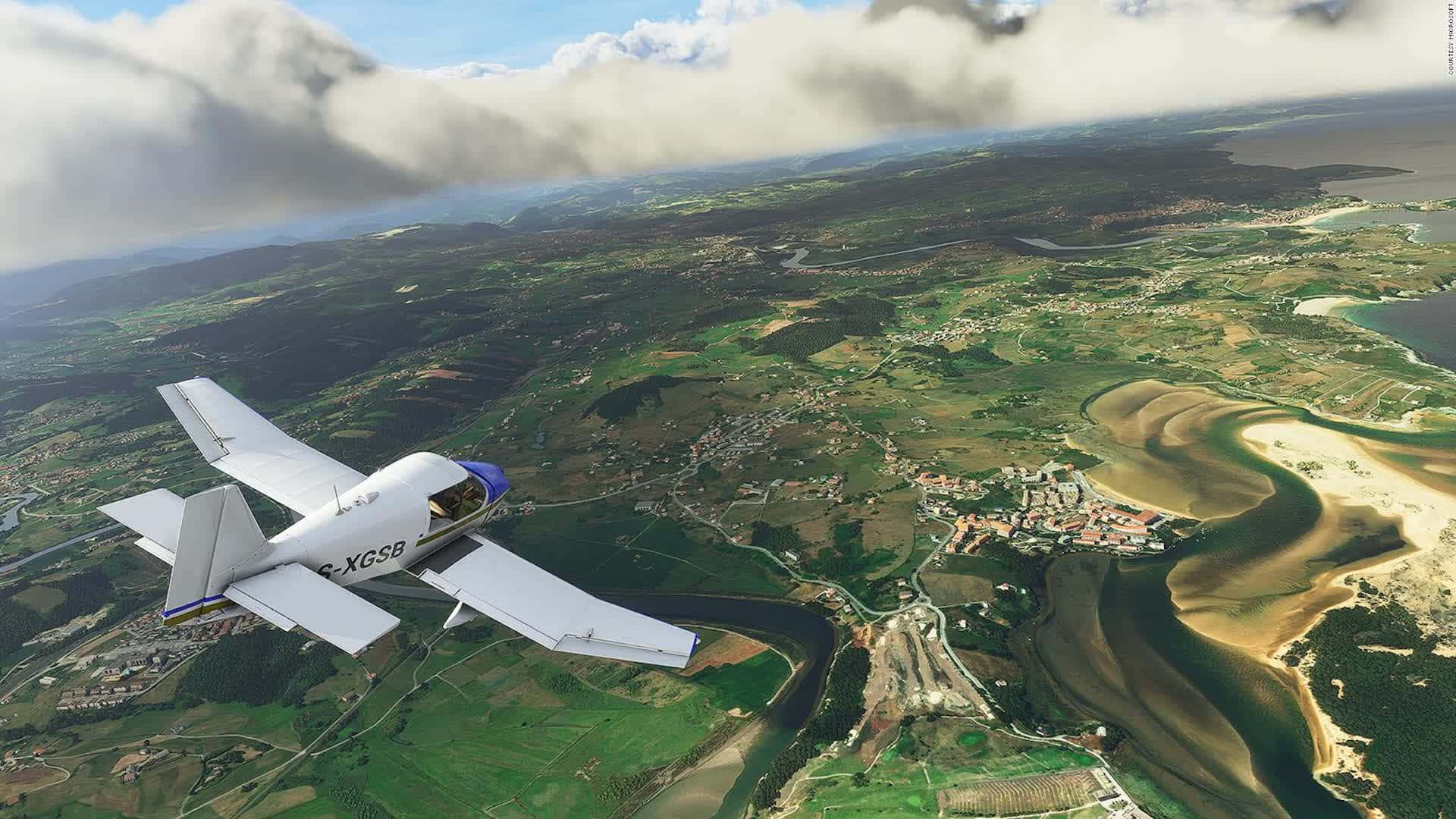 1080p Microsoft Flight Simulator 桌布 壁紙 — 一架白色單引擎飛機在部分多雲的天空下，翱翔於鬱鬱蔥蔥的綠色山谷之上，山谷中有蜿蜒的河流和沿海城鎮。
