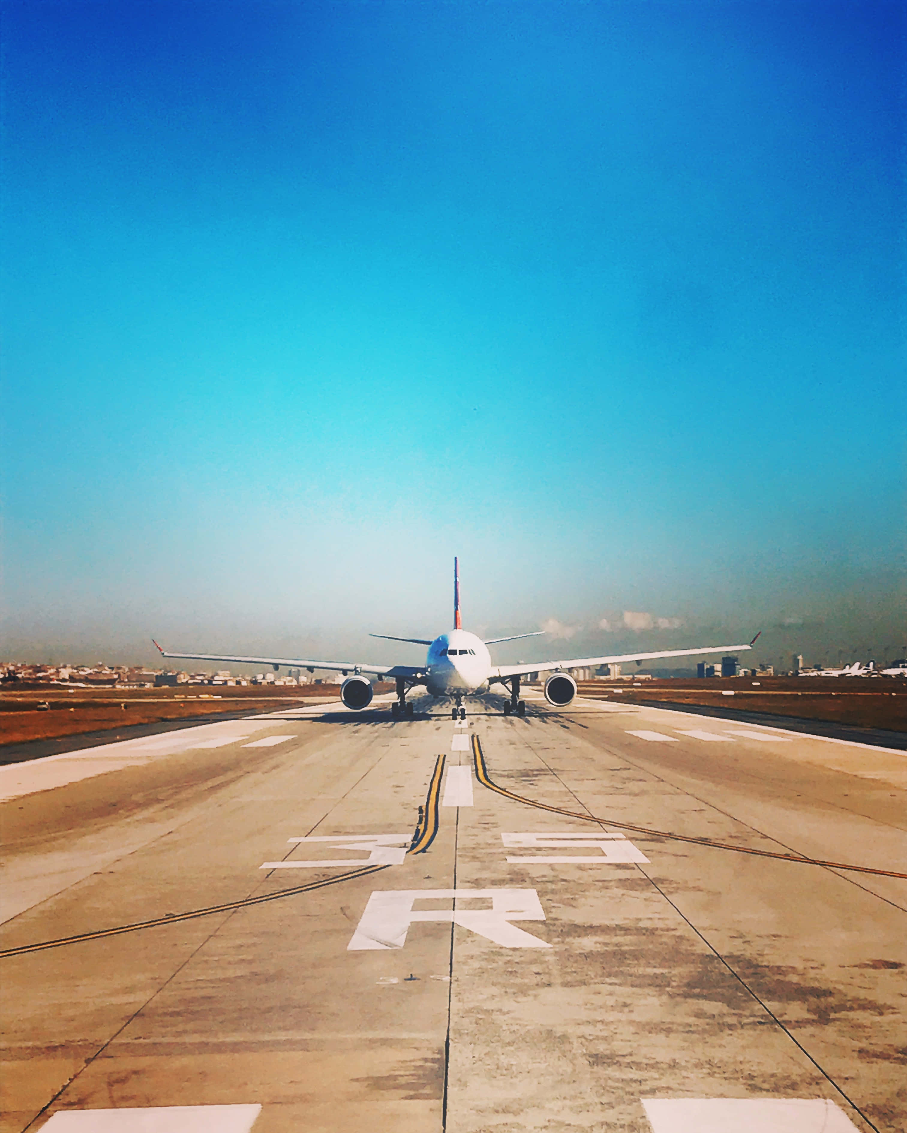 Runway 桌布 壁紙 — 一架白色客機停在陽光明媚的跑道上，背景是廣闊的藍天和遠處的城市景觀。