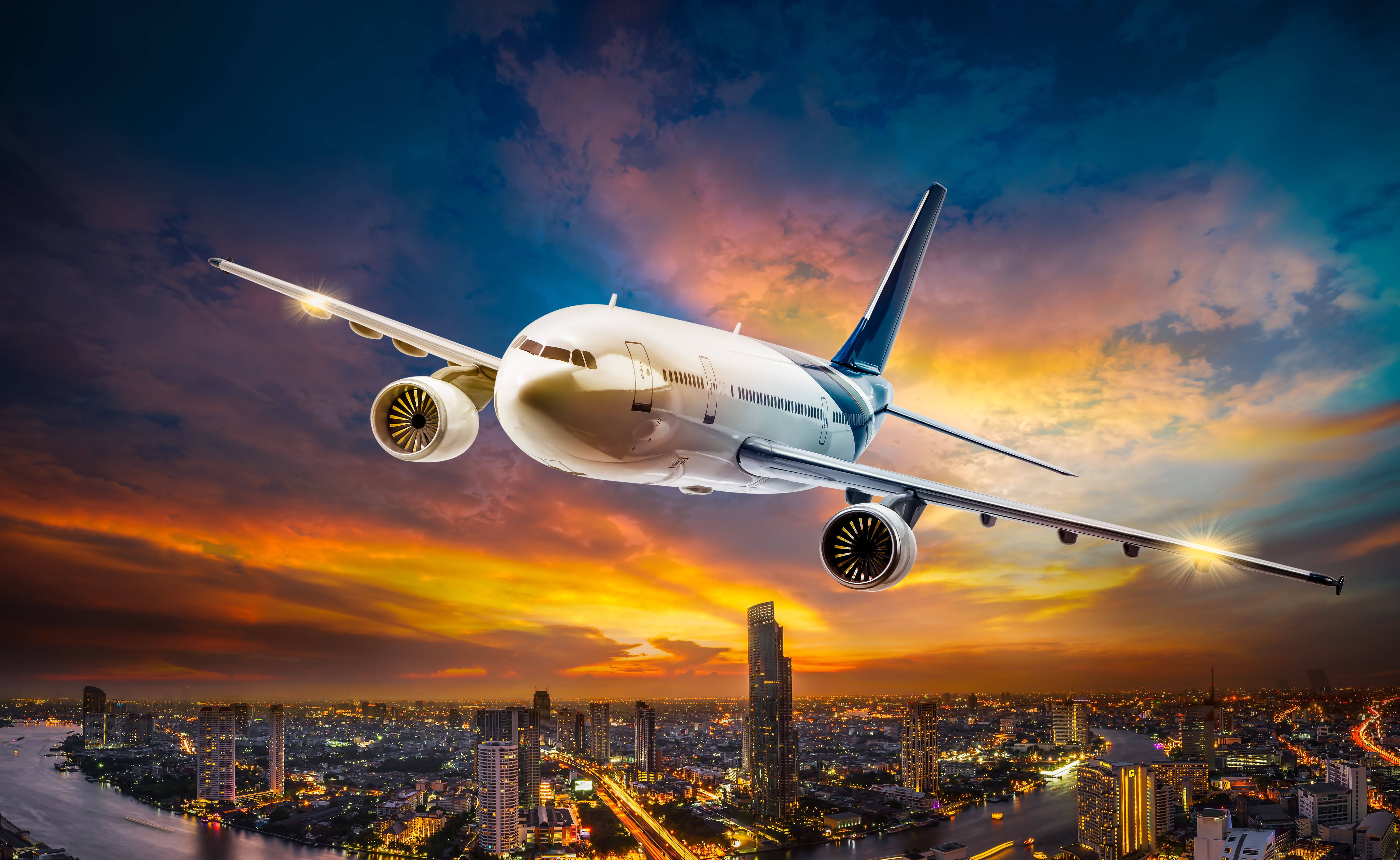 HD Plane 桌布 壁紙 — 一架白色客機在日落時分翱翔於發光的城市景觀之上，下方有火紅的橙色雲朵和閃爍的燈光。