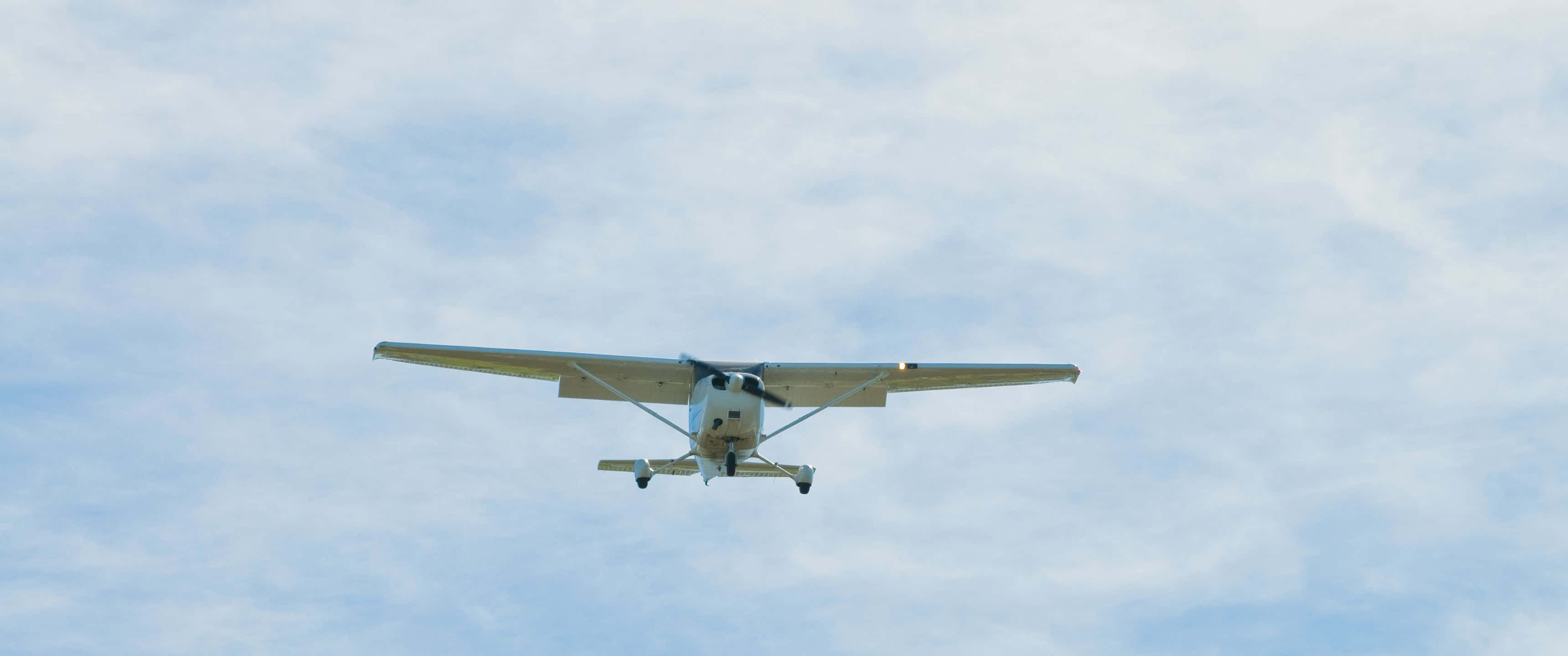 3440x1440p Small Planes 桌布 壁紙 — 一架白色小型螺旋槳飛機正面飛向帶有薄雲的柔和藍天。