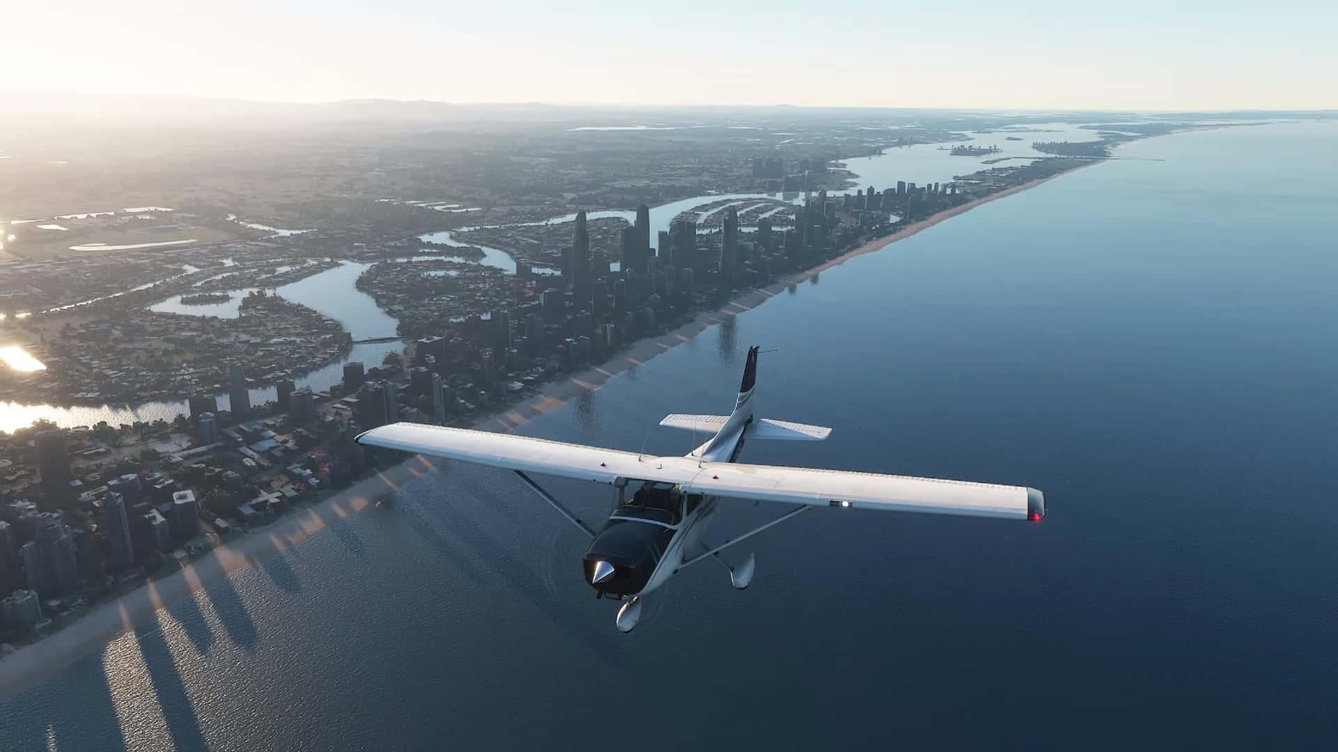 1920x1080 Microsoft Flight Simulator 桌布 壁紙 — 一架白色小飛機在日出時低飛過沿海城市景觀，長長的陰影在水面上延伸。