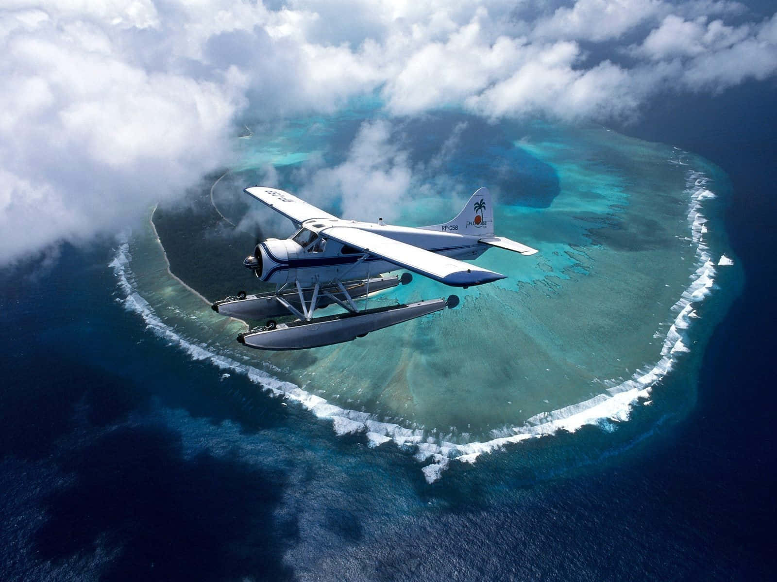 Helicopter 桌布 壁紙 — 一架白色水上飛機優雅地滑翔在一個被深藍色海洋和蓬鬆白雲環繞的碧綠環礁上空。