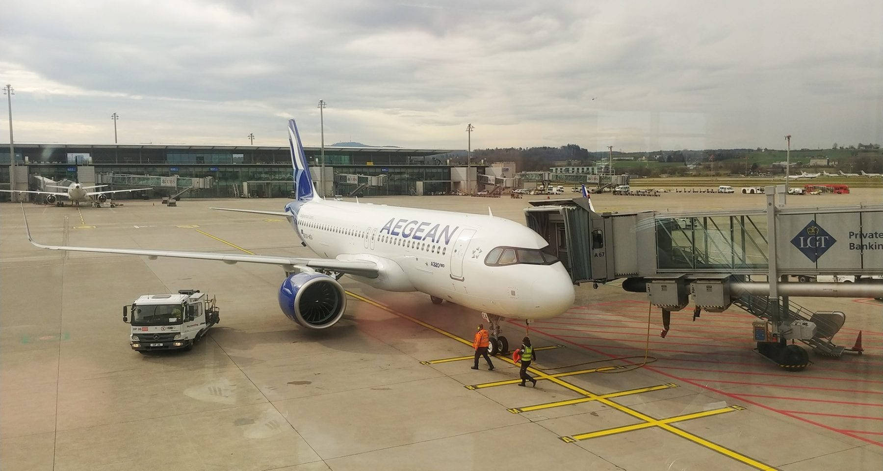 Airport 桌布 壁紙 — 一架白色的愛琴海航空飛機停泊在機場閘口，附近有地勤人員和登機橋，天空多雲。