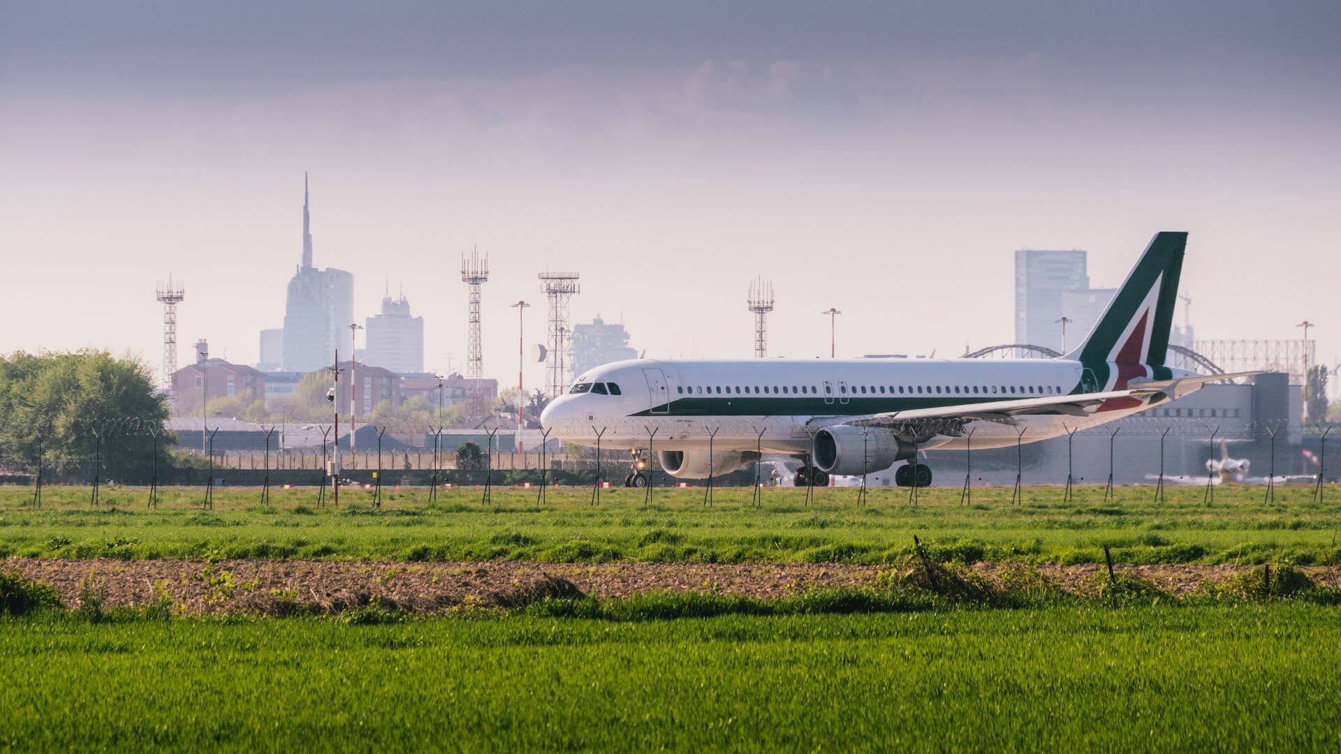 飛機 桌布 壁紙 — 一架白色的義大利航空飛機在跑道上滑行，前景是綠色田野，背景是朦朧的城市天際線。