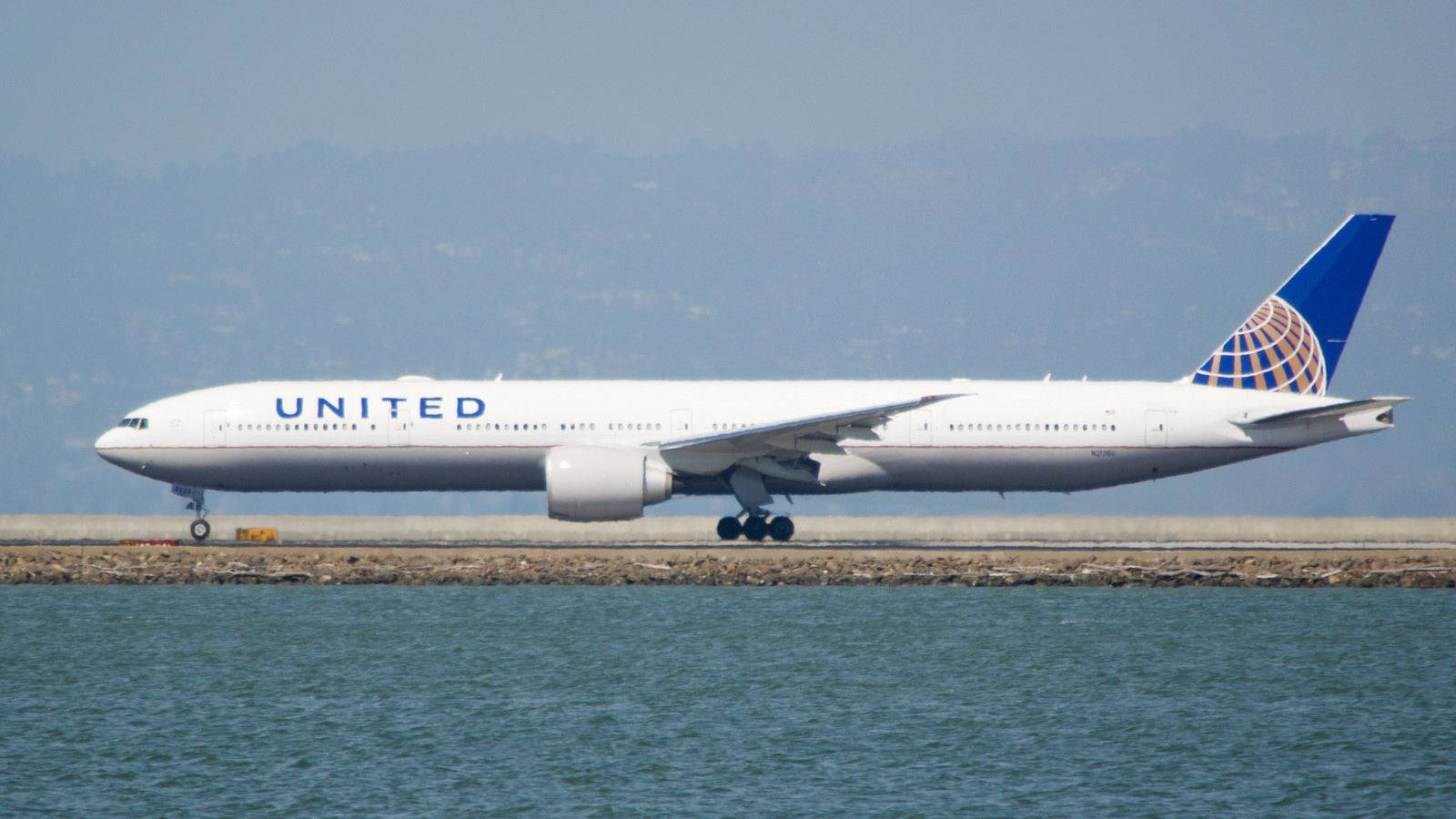 Plane 桌布 壁紙 — 一架白色的聯合航空波音777在平靜的藍色水邊的跑道上滑行，背景有朦朧的山丘。