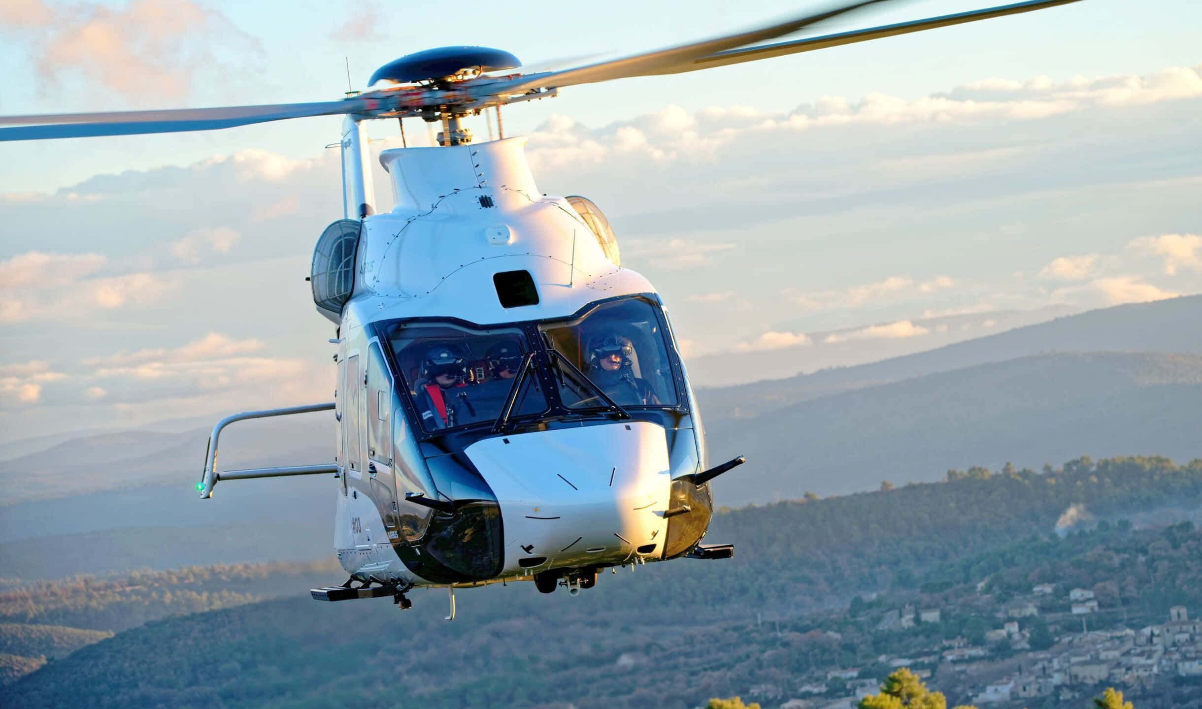 2440x1440 Helicopters 桌布 壁紙 — 一架白色直升機在陽光明媚的山區景觀上空盤旋，下方散落著房屋。