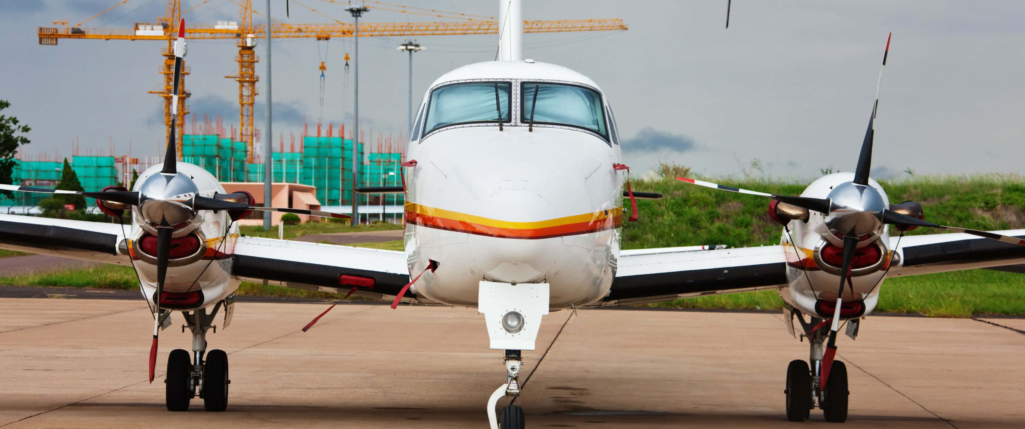 3440x1440p Small Planes 桌布 壁紙 — 一架白色雙引擎螺旋槳飛機停在停機坪上，背景可見建築起重機和綠色鷹架。