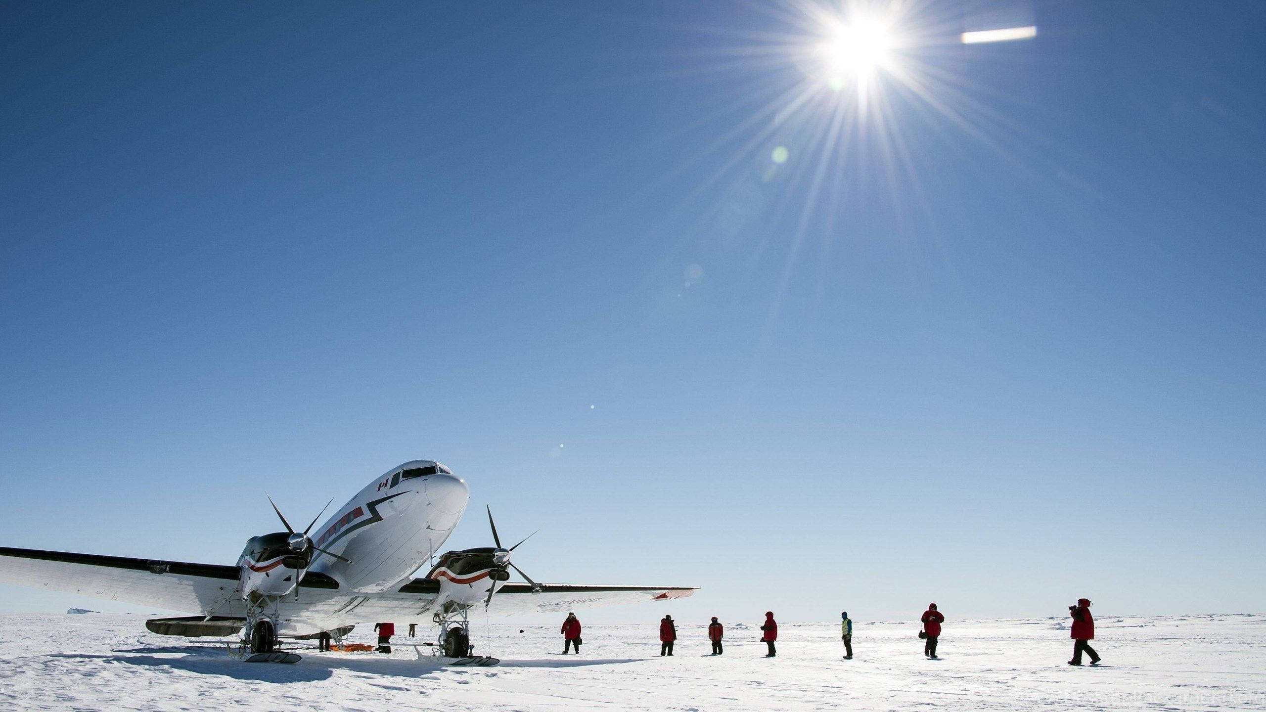 Antarctica 桌布 壁紙 — 一架白色雙引擎飛機在無盡的雪地上停歇，烈日當空，身穿紅色派克大衣的人影在冰凍的苔原上行走。