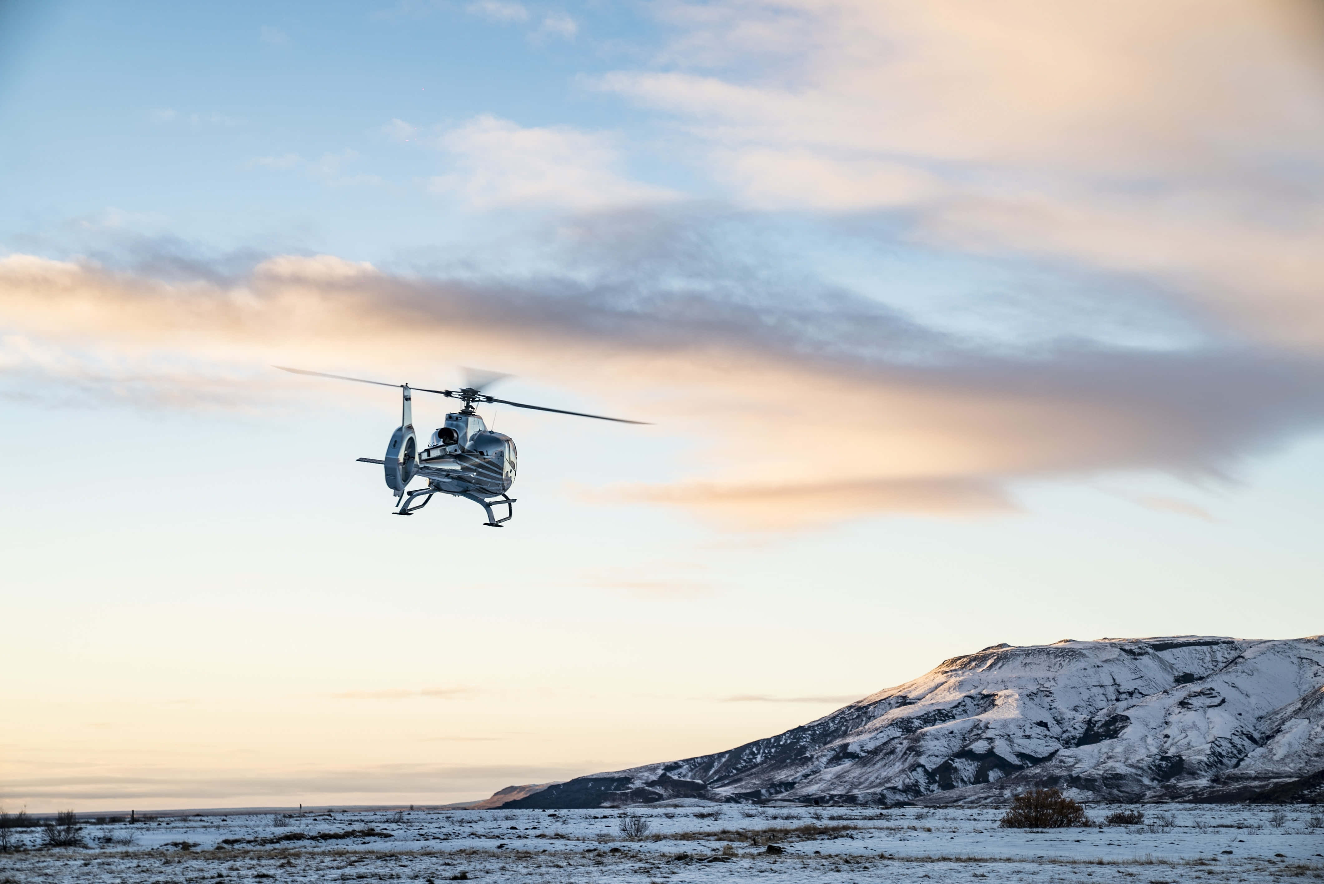 4K Helicopters 桌布 壁紙 — 一架直升機在日落時分盤旋在積雪的山脈上空，伴隨著柔和的粉彩雲朵。