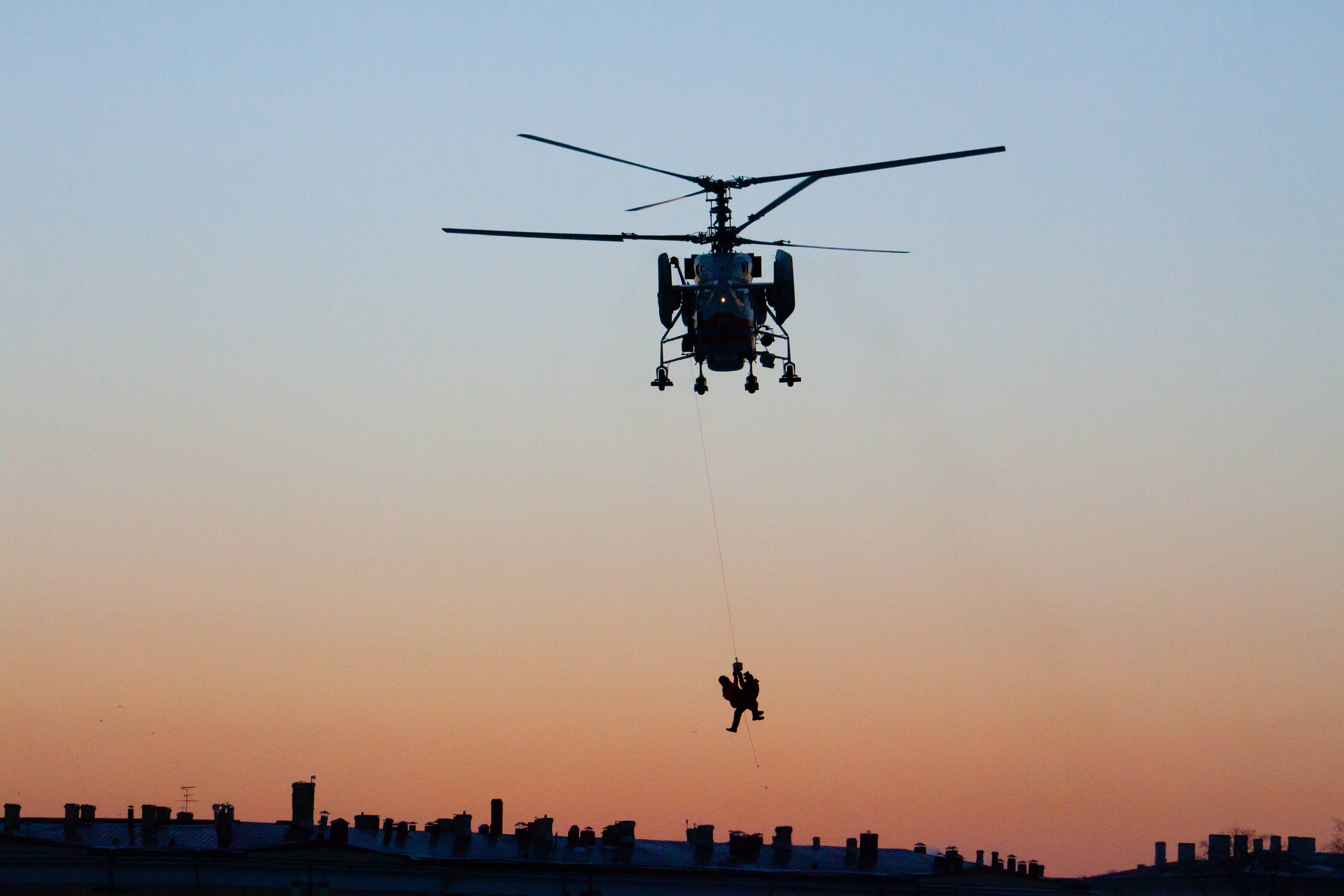 helicopter 4K 桌布 壁紙 — 一架直升機在暮色天空的襯托下，用繩索將一個人降落到城市天際線上空。