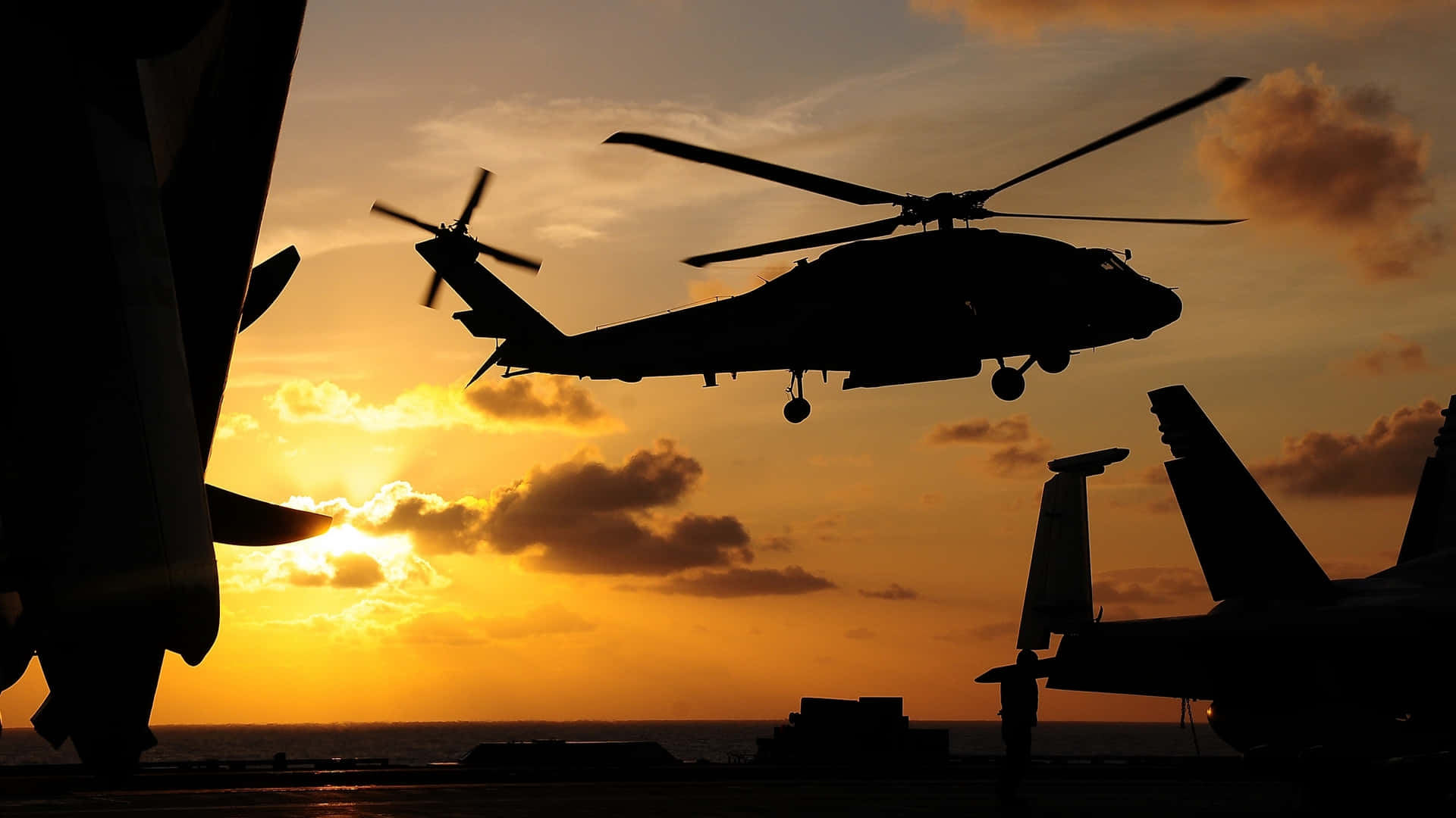 Helicopters 桌布 壁紙 — 一架直升機在熾熱的夕陽映襯下，盤旋在航空母艦甲板上方，旁邊還有其他飛機。