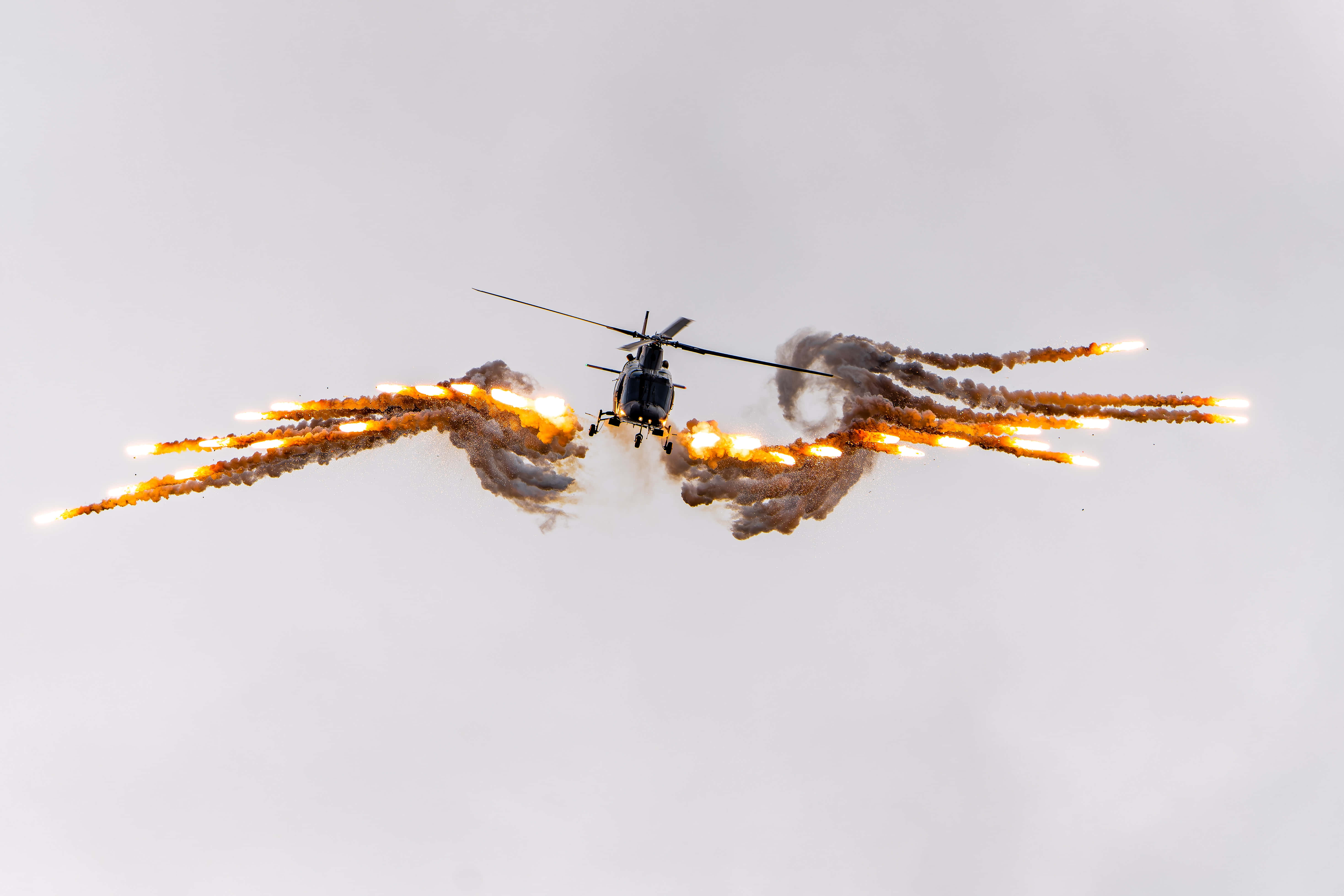 Helicopter 桌布 壁紙 — 一架直升機在蒼白的天空下低飛，拖曳著對稱爆發的熾熱橙色信號彈。