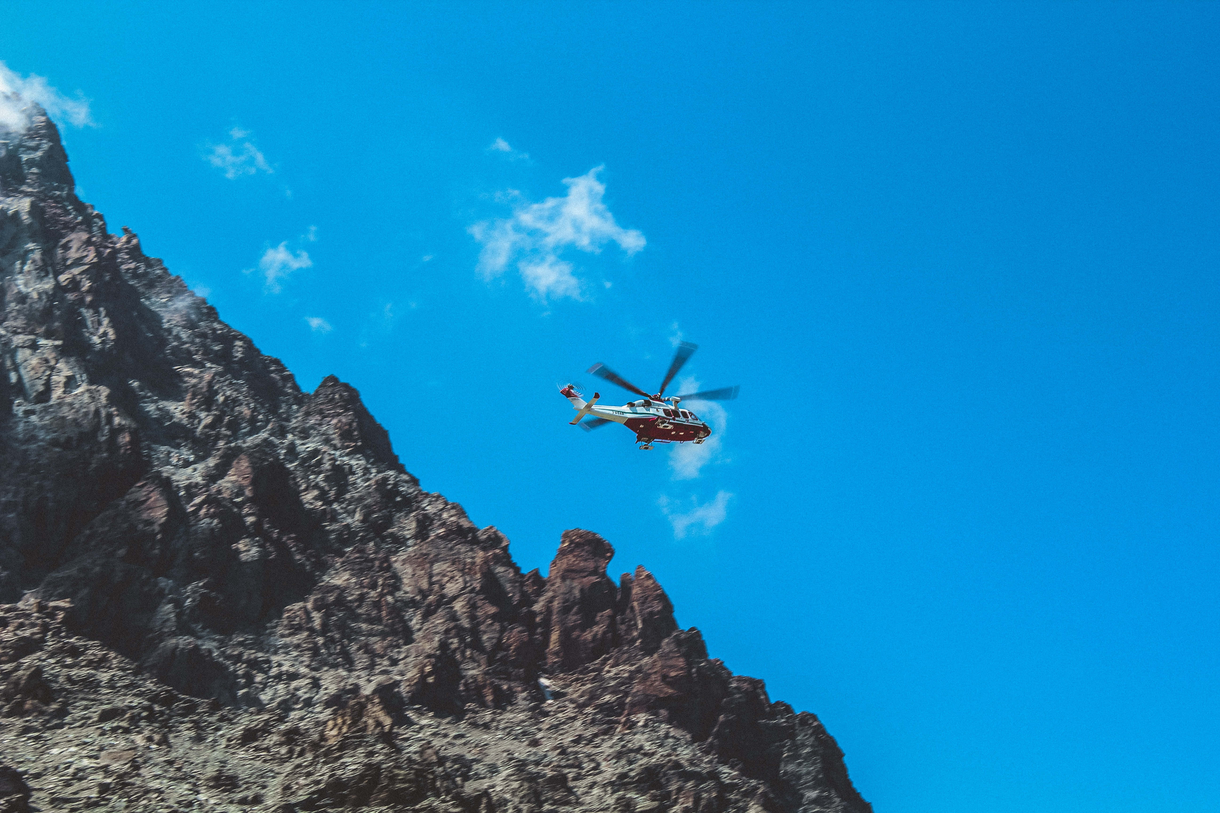 Helicopter 桌布 壁紙 — 一架直升機在蔚藍的天空中翱翔，飛越崎嶇岩石山峰，天空中有散落的雲朵。