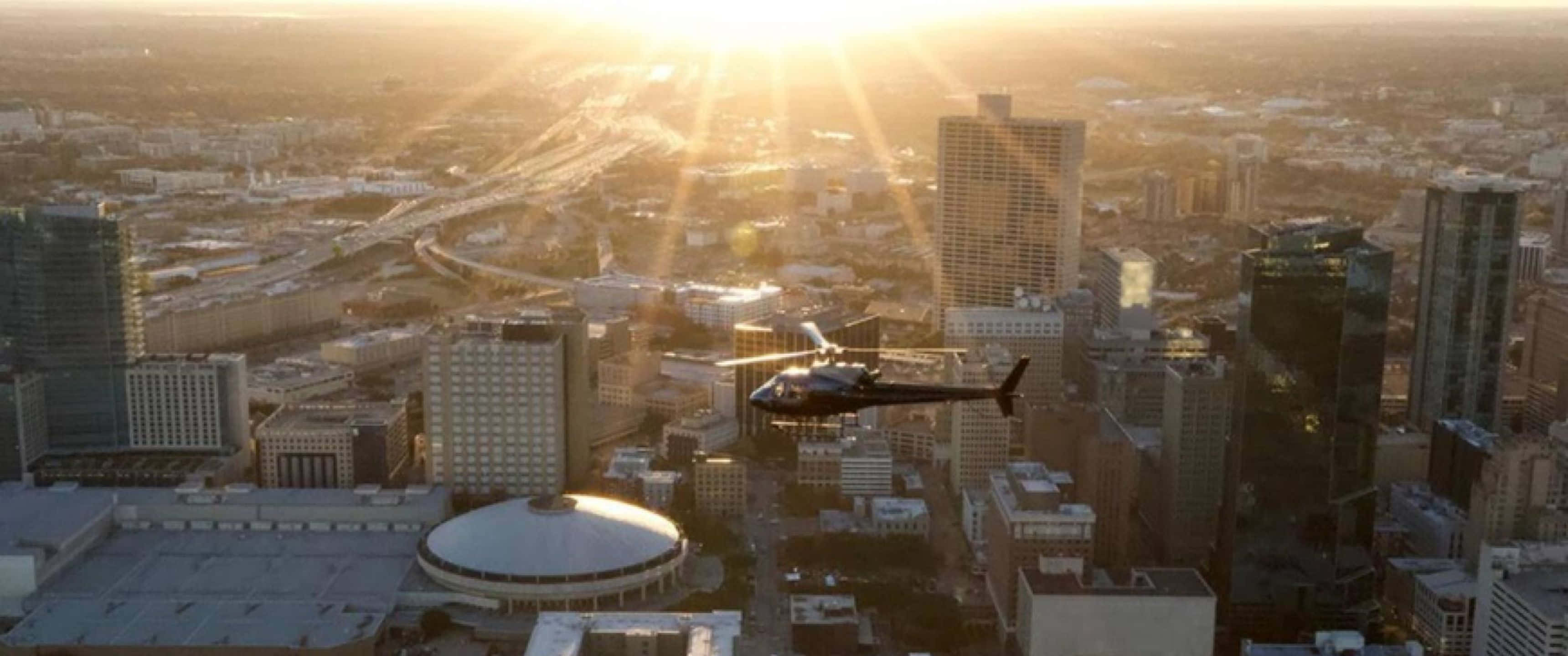 3440x1440p Helicopters 桌布 壁紙 — 一架直升機在金色日落時分飛越廣闊的城市景觀，燦爛的陽光穿過摩天大樓。