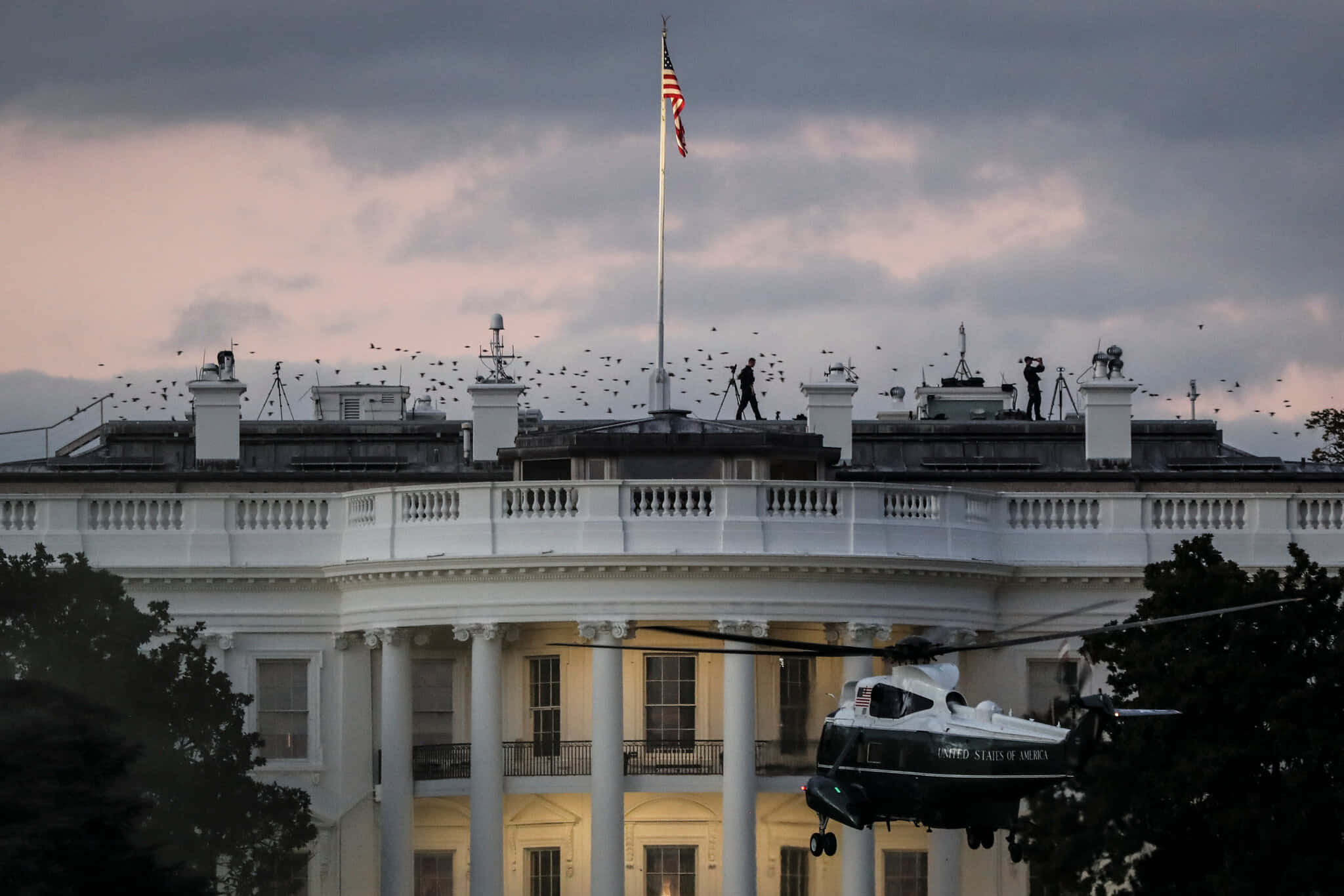white house 桌布 壁紙 — 一架直升機在黃昏的天空下盤旋在白宮附近，鳥兒在屋頂上散開。
