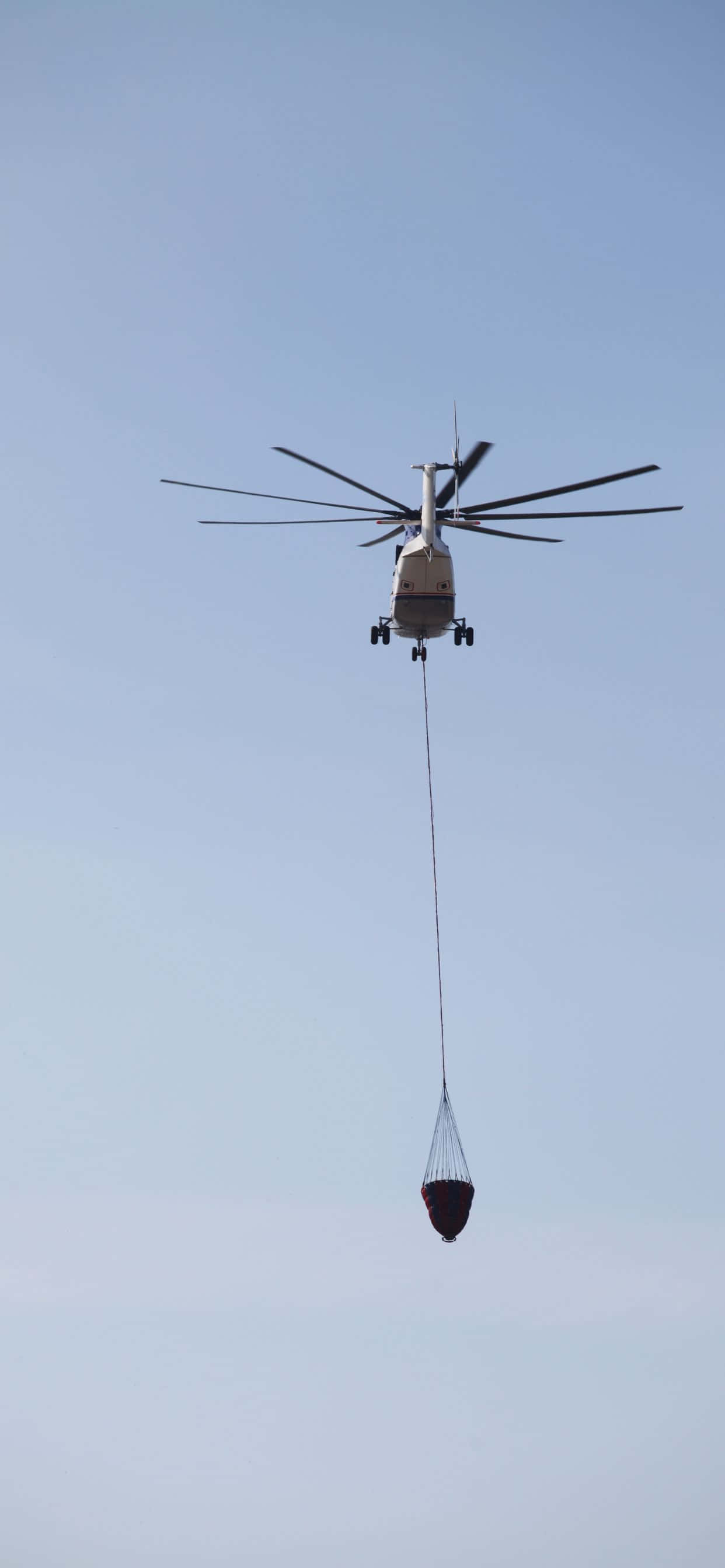 iPhone Xs Max Helicopters 桌布 壁紙 — 一架直升機懸掛在清澈的藍天中，用長繩放下一個紅色水桶。