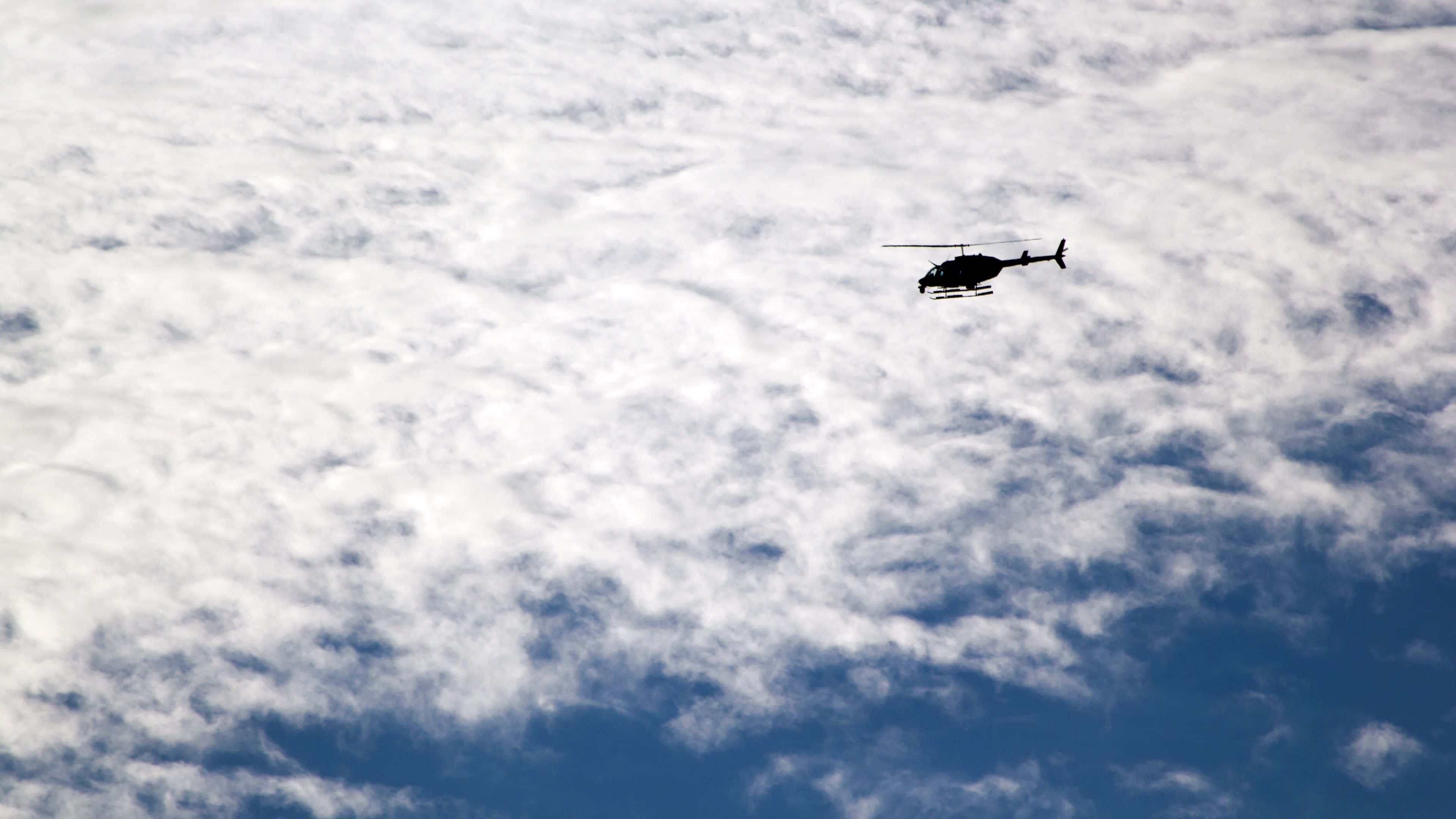 4K Helicopters 桌布 壁紙 — 一架直升機的剪影在深藍色天空的背景下，翱翔於質感豐富的白雲毯之上。