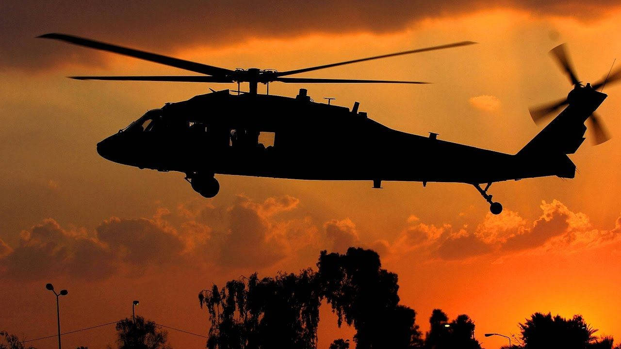 Helicopter 桌布 壁紙 — 一架直升機的剪影在火紅的橙色日落天空下翱翔，下方有戲劇性的雲彩和樹木輪廓。
