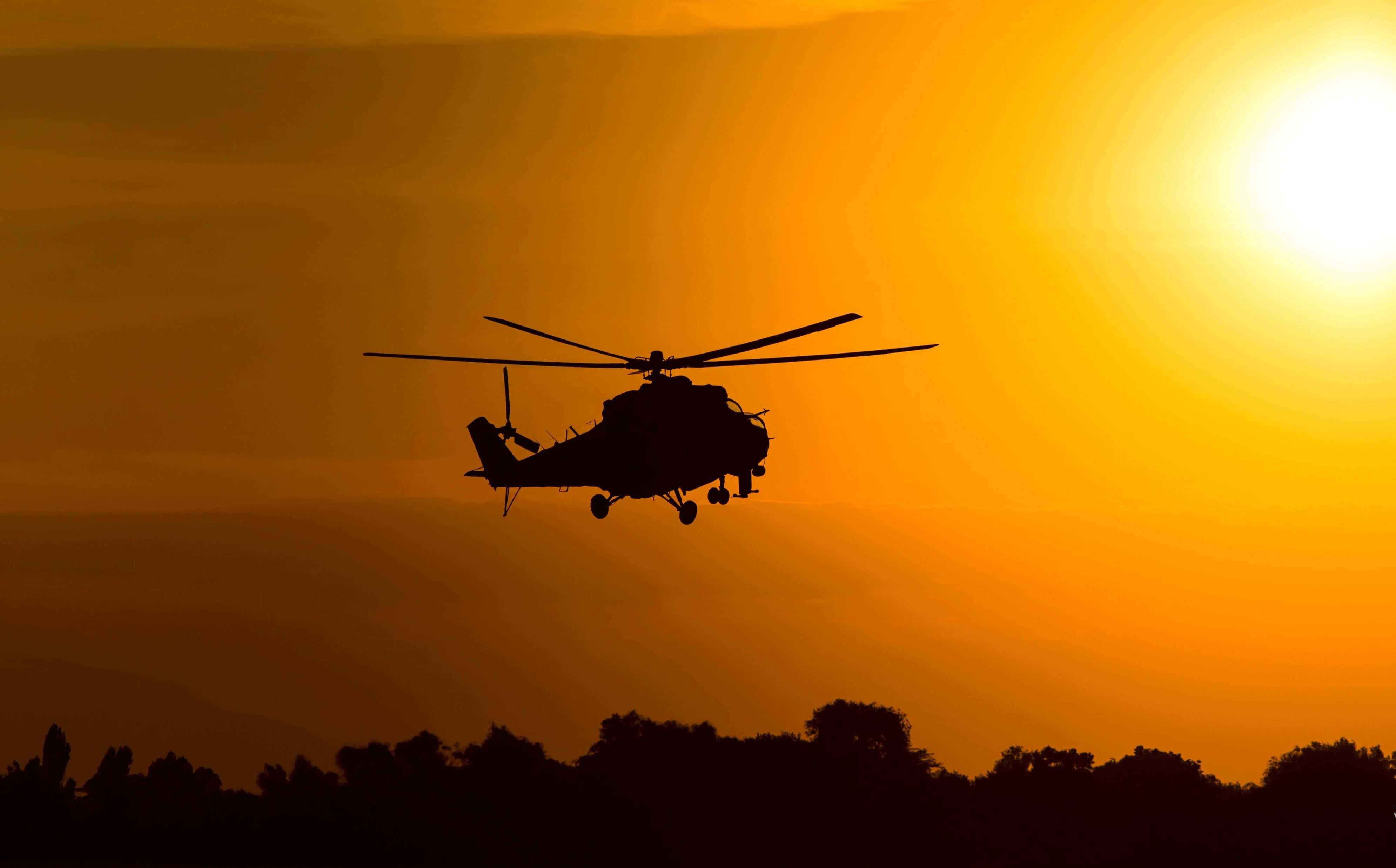 4K Helicopters 桌布 壁紙 — 一架直升機的剪影在黑暗的樹線上方，翱翔於火紅橙色的日落天空。