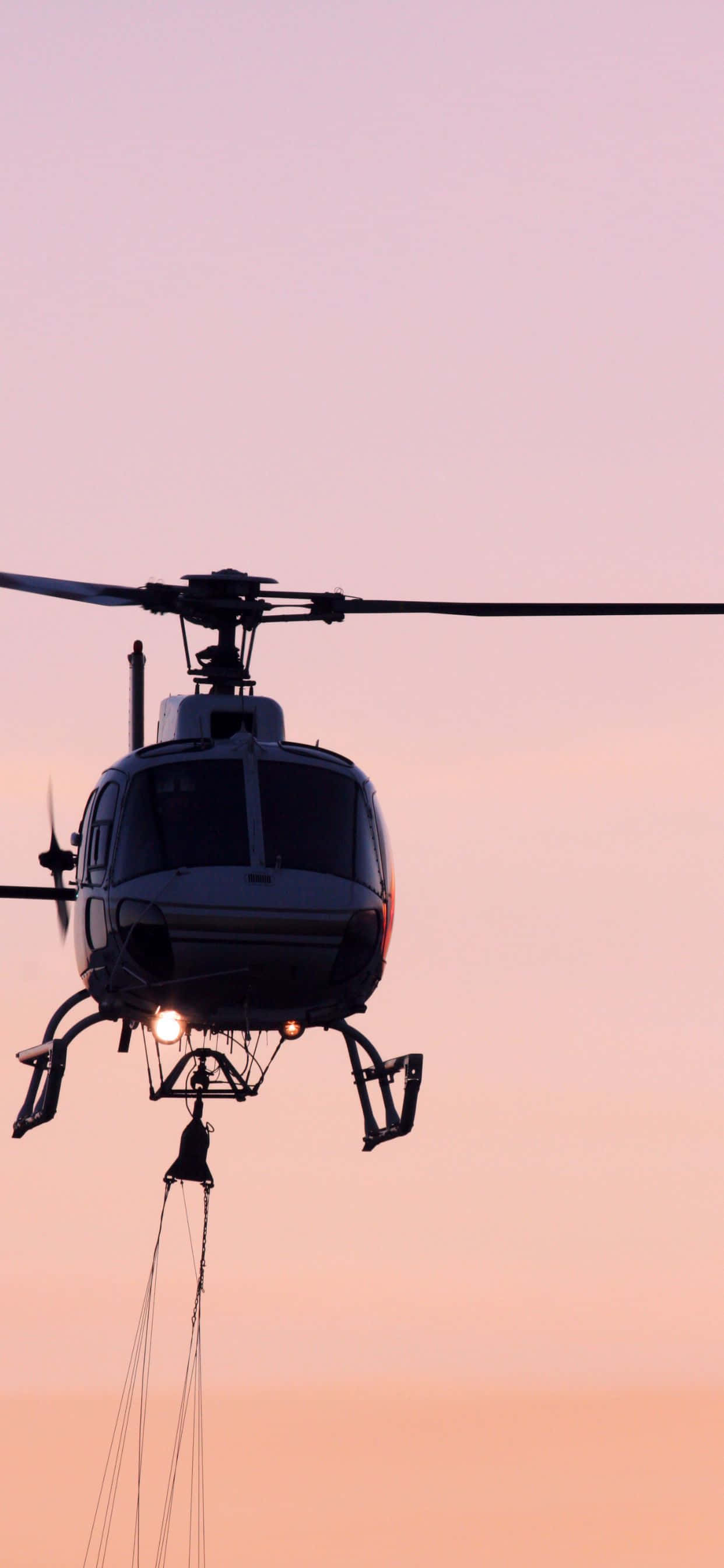 iPhone Xs Max Helicopters 桌布 壁紙 — 一架直升機的剪影懸掛在柔和的粉紅色和橙色暮色天空上，下方有發光的光線。