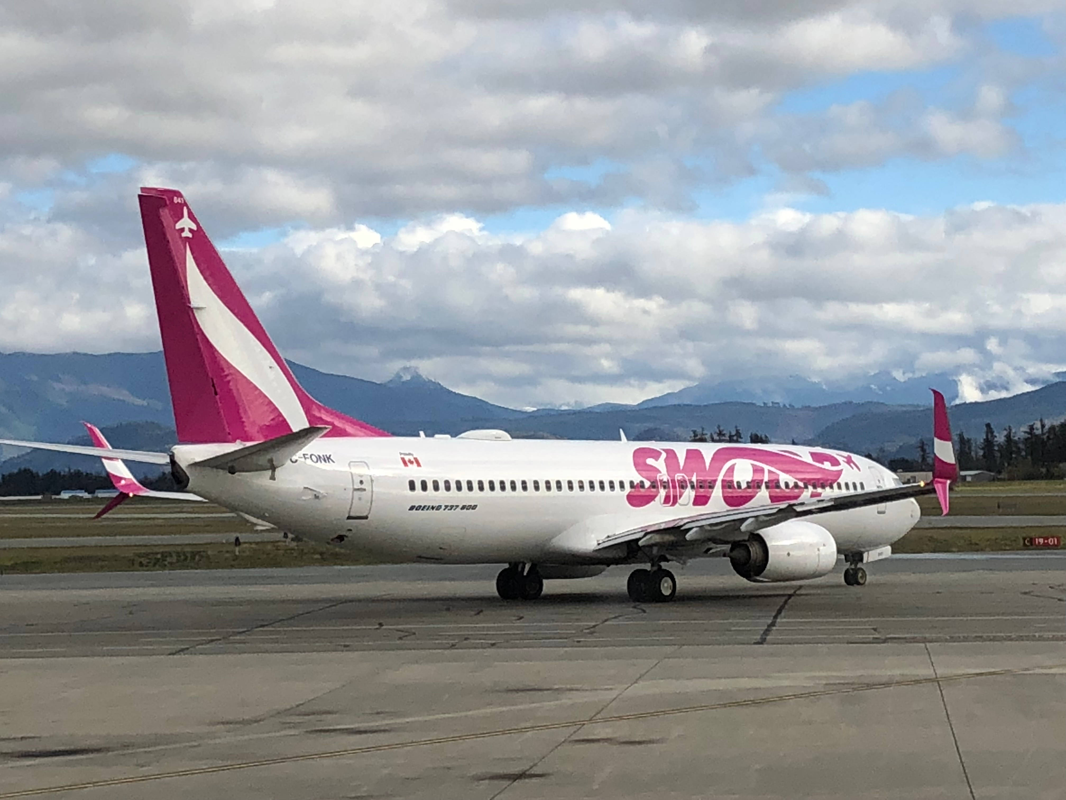 Aviation 桌布 壁紙 — 一架粉紅色和白色的Swoop Airlines波音737停放在停機坪上，背景是白雪覆蓋的山脈和多雲的天空。