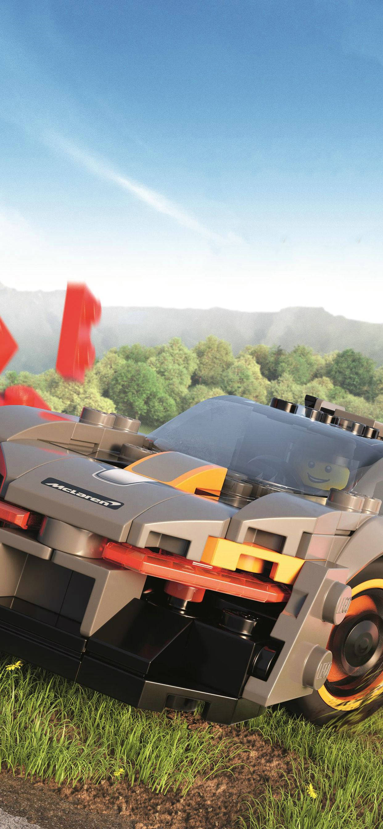 iPhone 桌布 壁紙 — 一架精細的LEGO McLaren跑車停放在草地上，背景是山脈和樹木，天空清澈湛藍。