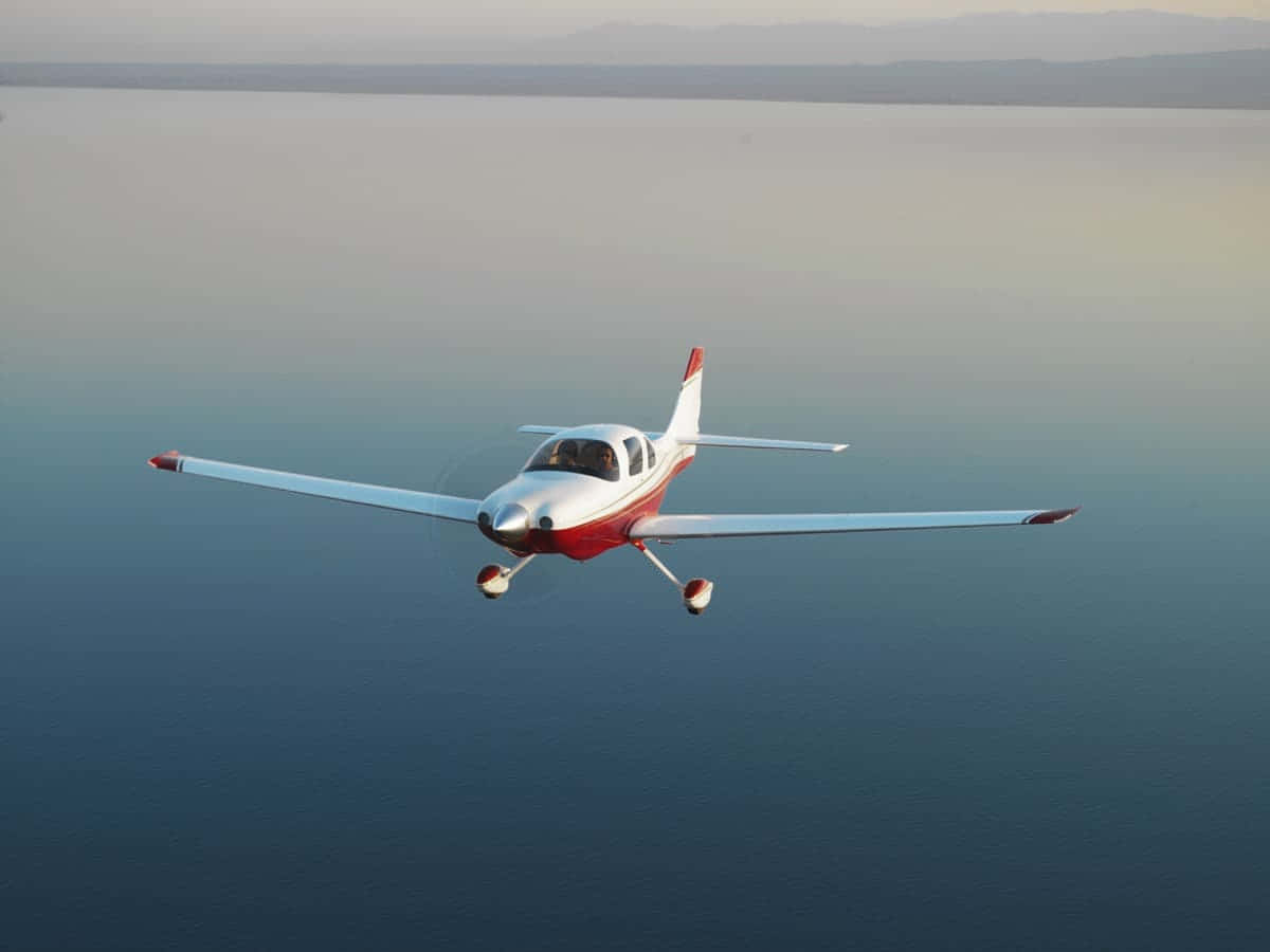 Helicopter 桌布 壁紙 — 一架紅白單引擎飛機低飛過平靜的藍色水面，遠山在柔和的金色天空下。
