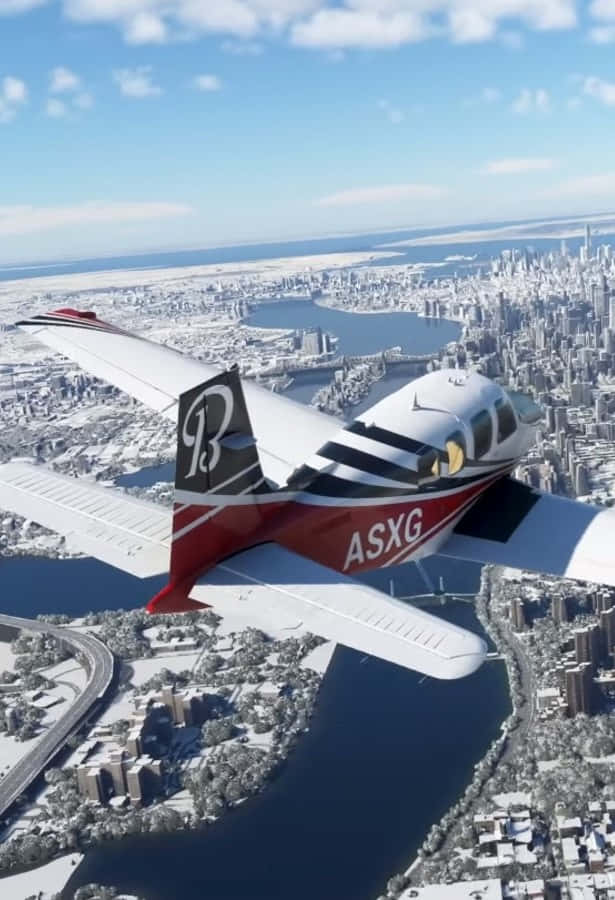 Android Microsoft Flight Simulator 桌布 壁紙 — 一架紅白單引擎飛機在明亮的藍天下低飛過被雪覆蓋的城市景觀，下方有河流和橋樑。