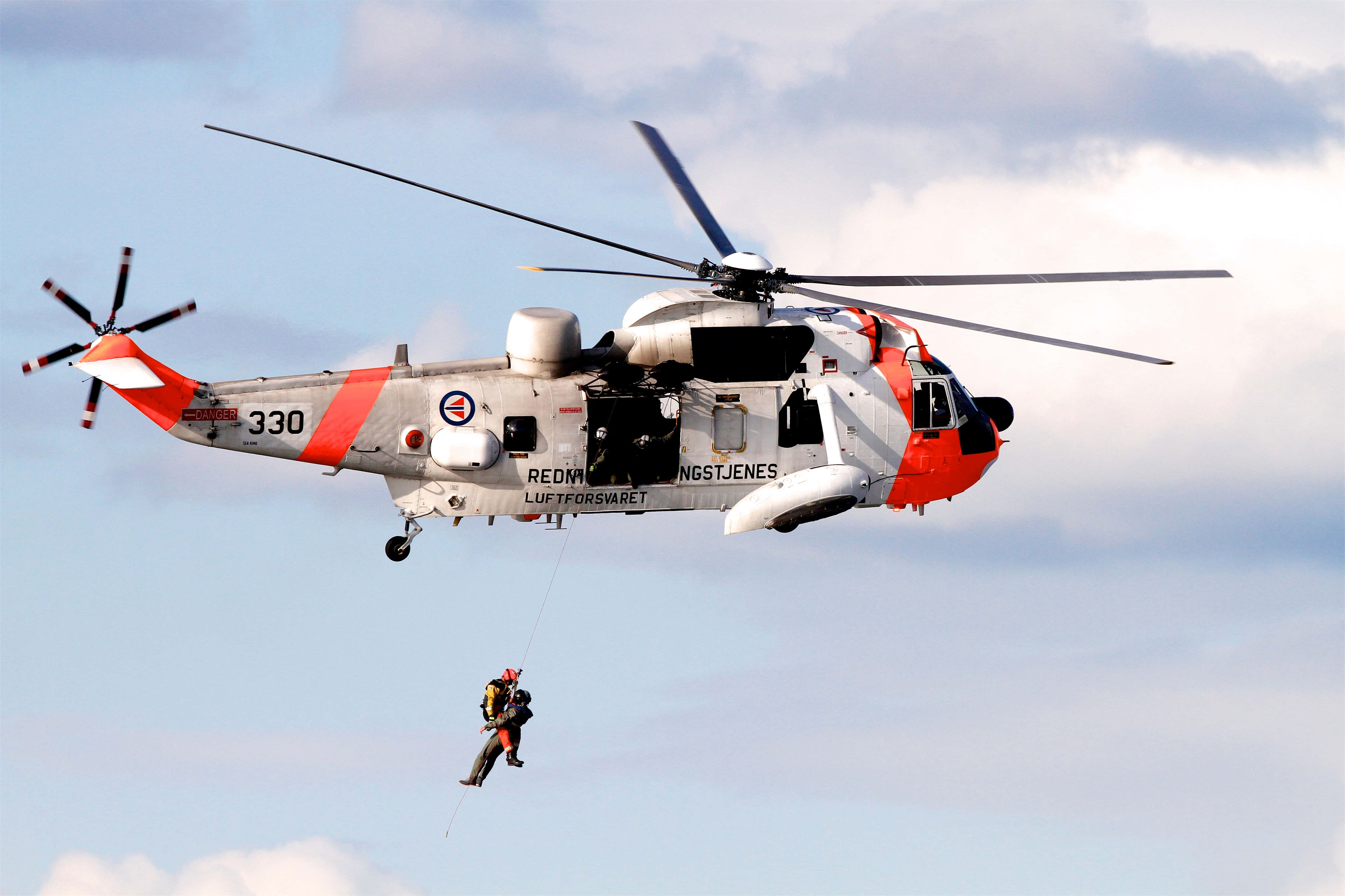 Helicopter 桌布 壁紙 — 一架紅白塗裝的挪威救援直升機在半空中盤旋，透過繩索將救援人員降下，背景是陰天。