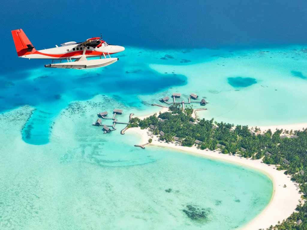 Maldives Island 桌布 壁紙 — 一架紅白相間的水上飛機在碧綠的海水上翱翔，飛越一個擁有水上平房和白色沙灘的熱帶島嶼度假村。