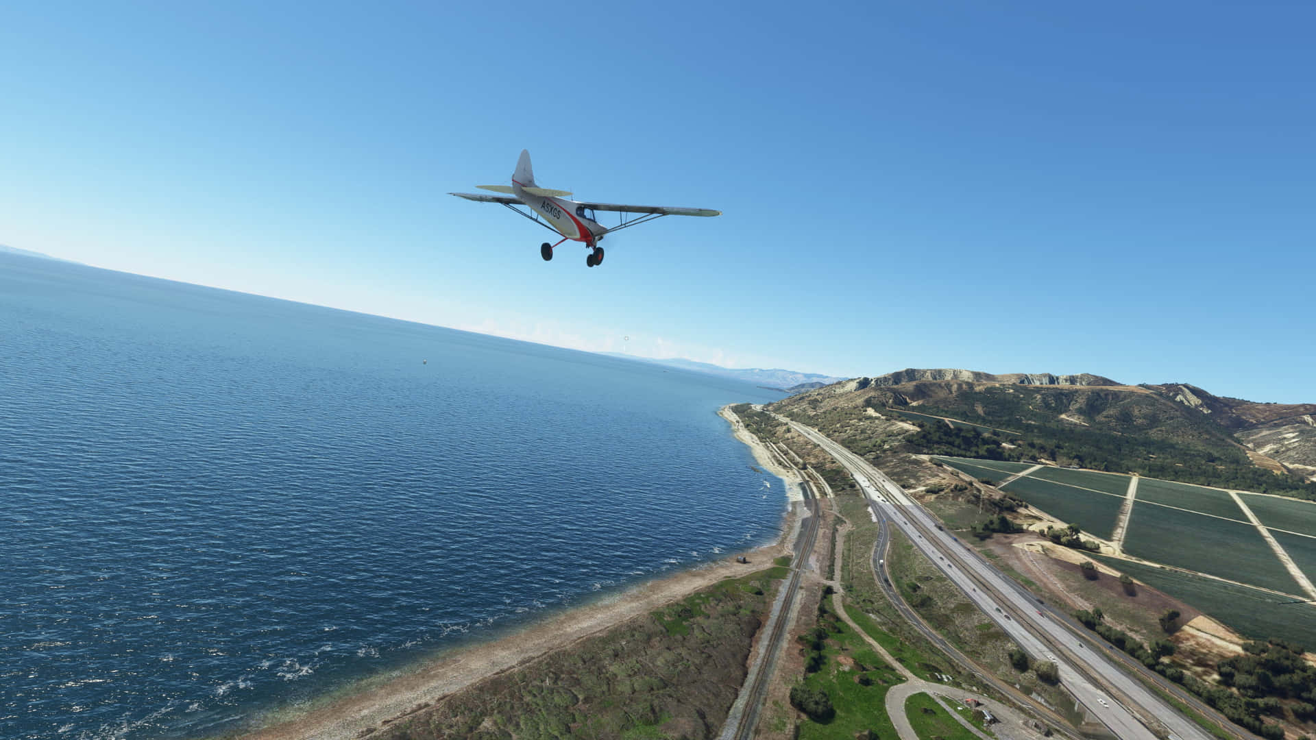 1920x1080 Microsoft Flight Simulator 桌布 壁紙 — 一架紅白色的小型飛機在晴朗藍天下，低飛過沿海高速公路，下方是藍色海洋和綠色山丘。