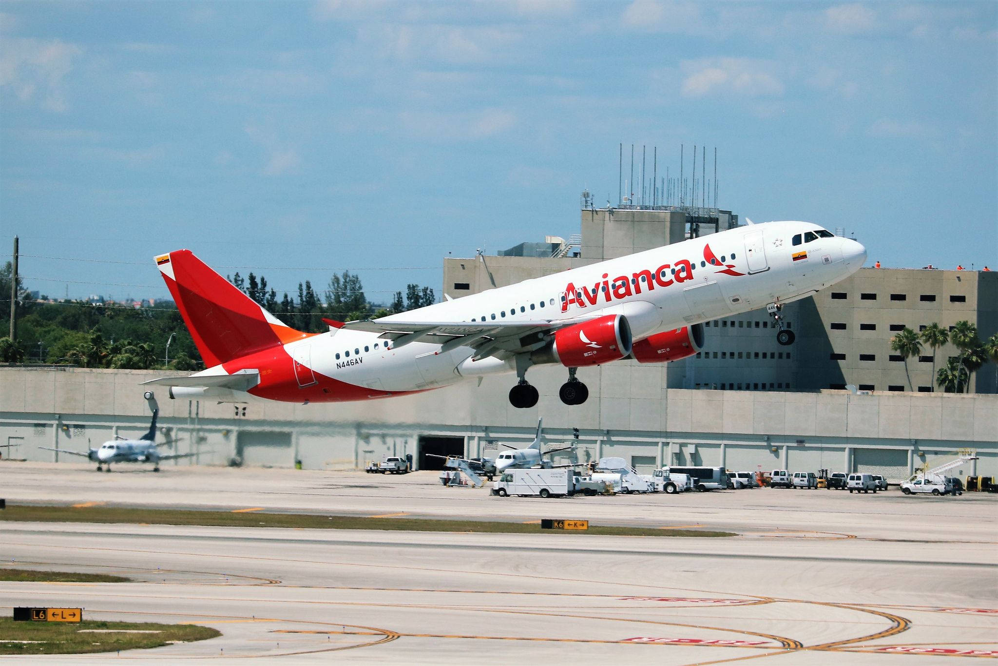 Airport 桌布 壁紙 — 一架紅色尾翼的 Avianca 飛機從陽光普照的機場跑道起飛，周圍是航廈和地面車輛。