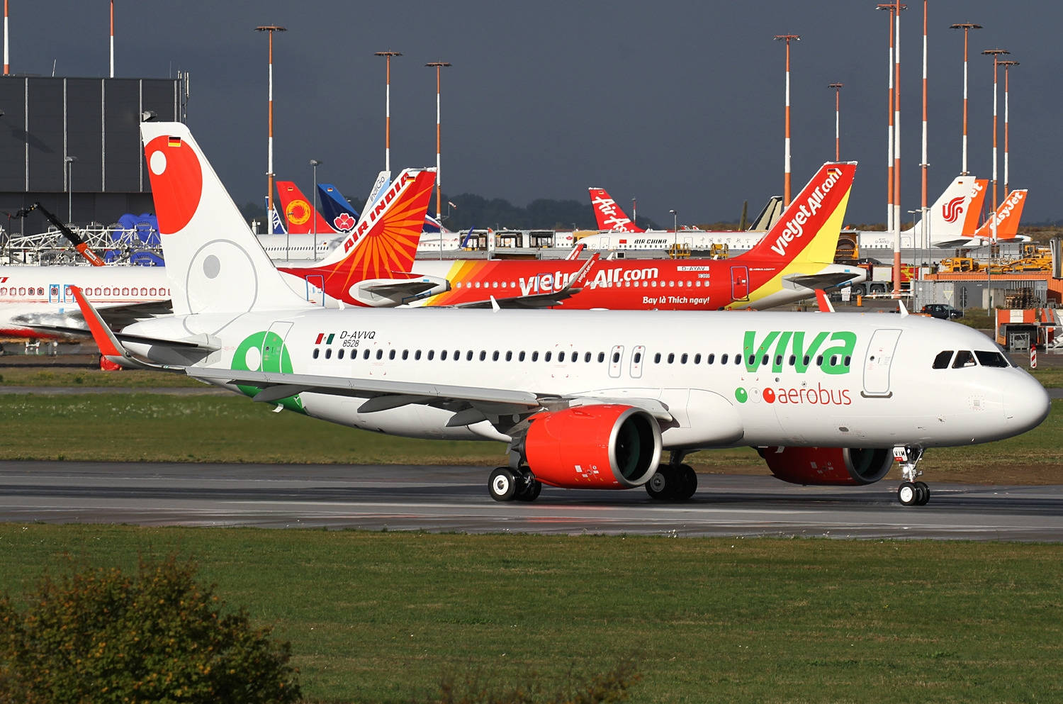 飛機 桌布 壁紙 — 一架紅色引擎的Viva Aerobus飛機在雲層密布的天空下滑行，背景有其他色彩繽紛的飛機。