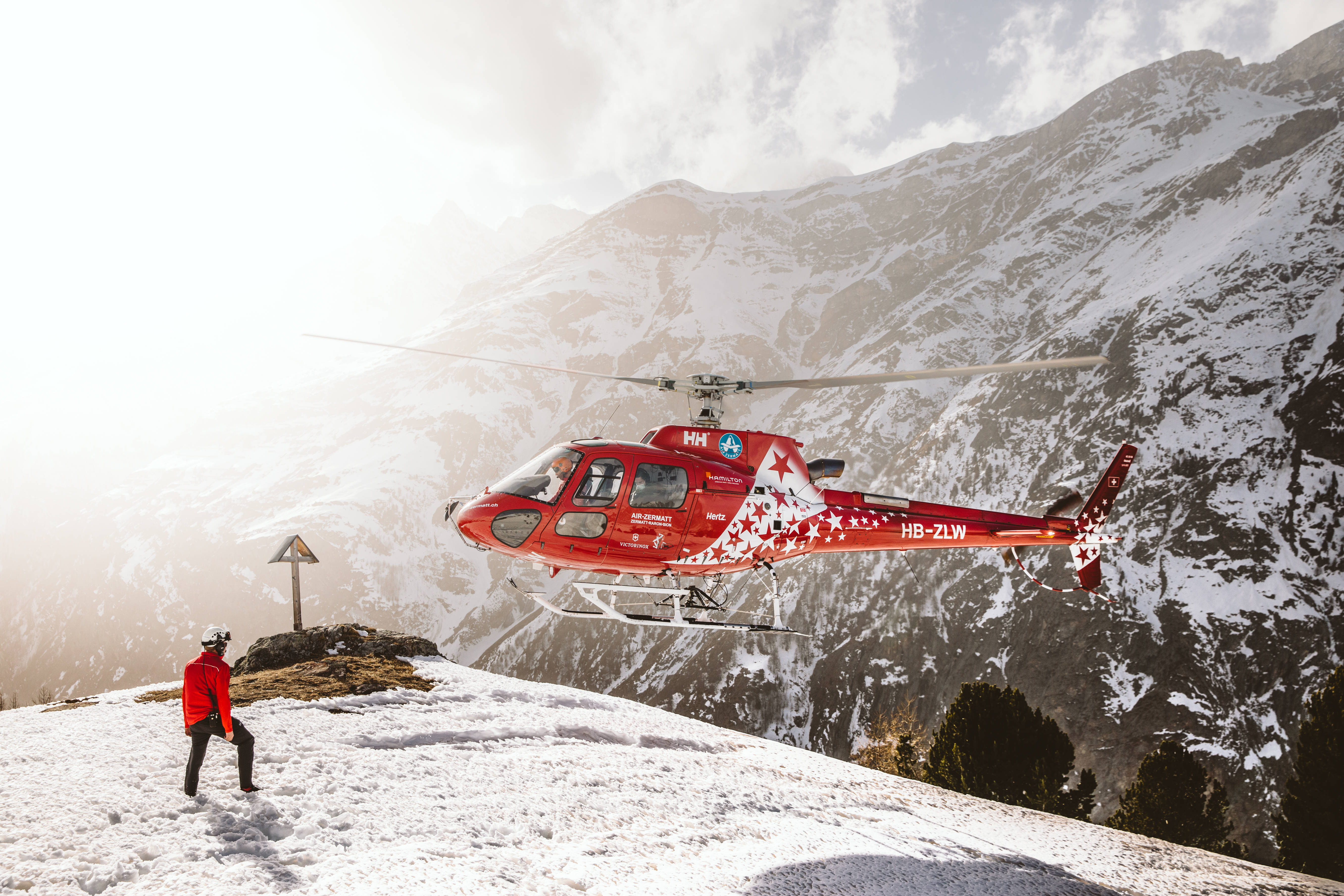 Helicopter 桌布 壁紙 — 一架紅色救援直升機在白雪覆蓋的山頂上空盤旋，一名身穿紅色外套的人站在下方。