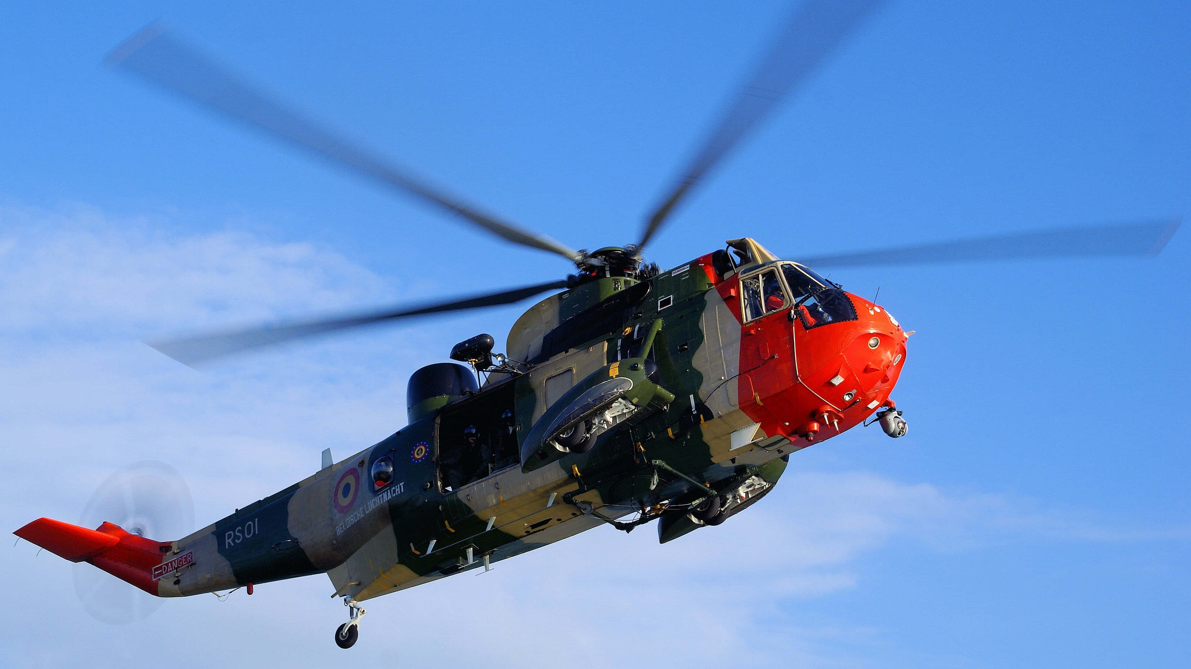 4K Helicopters 桌布 壁紙 — 一架紅色機頭、迷彩塗裝的軍用直升機在晴朗的藍天下飛行，旋轉的槳葉帶有動態模糊。